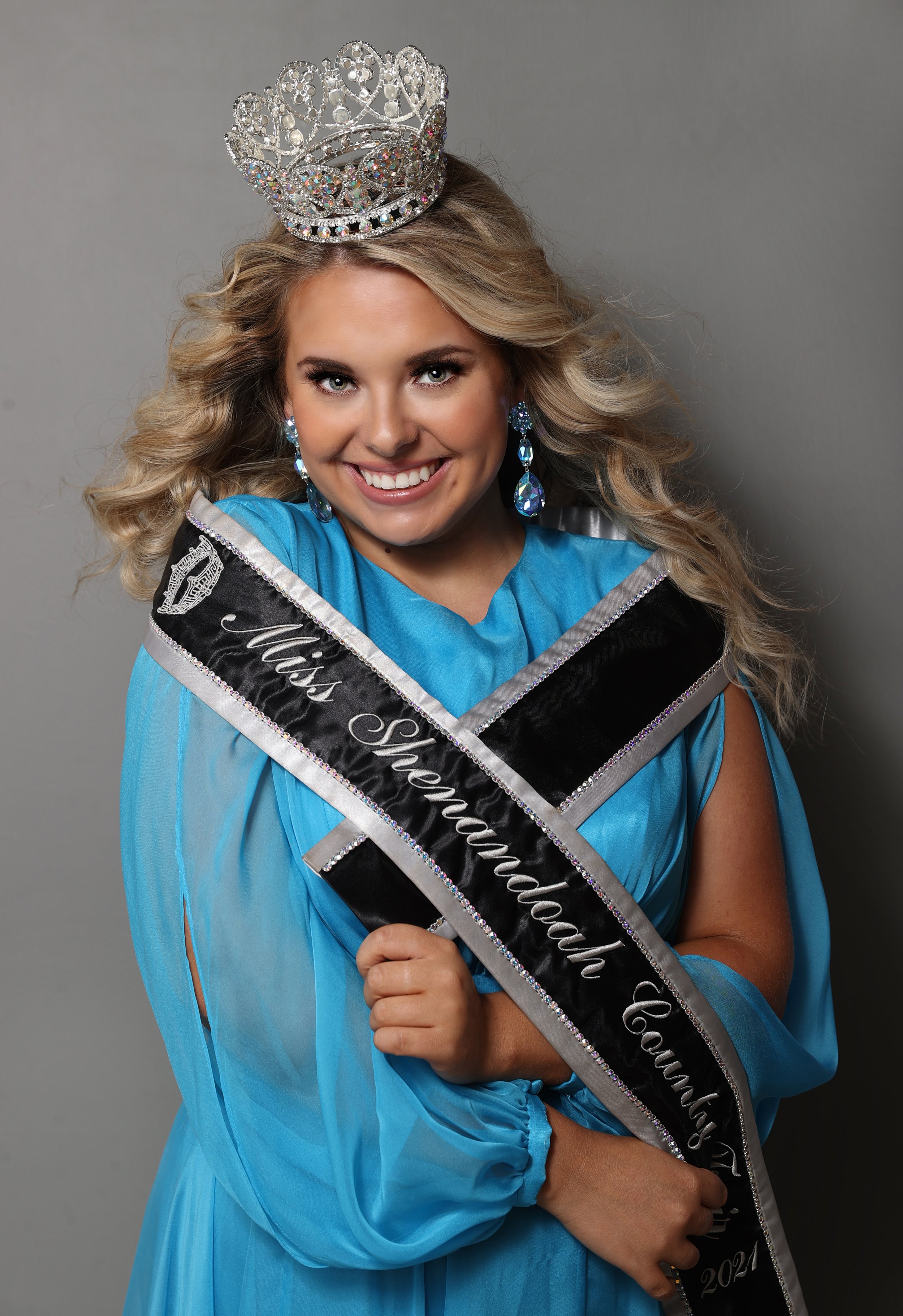 A woman with blonde curly hair wearing a crown, shining earrings, and a blue dress, holding a sash that reads 'Miss Shepardland County 2021.'