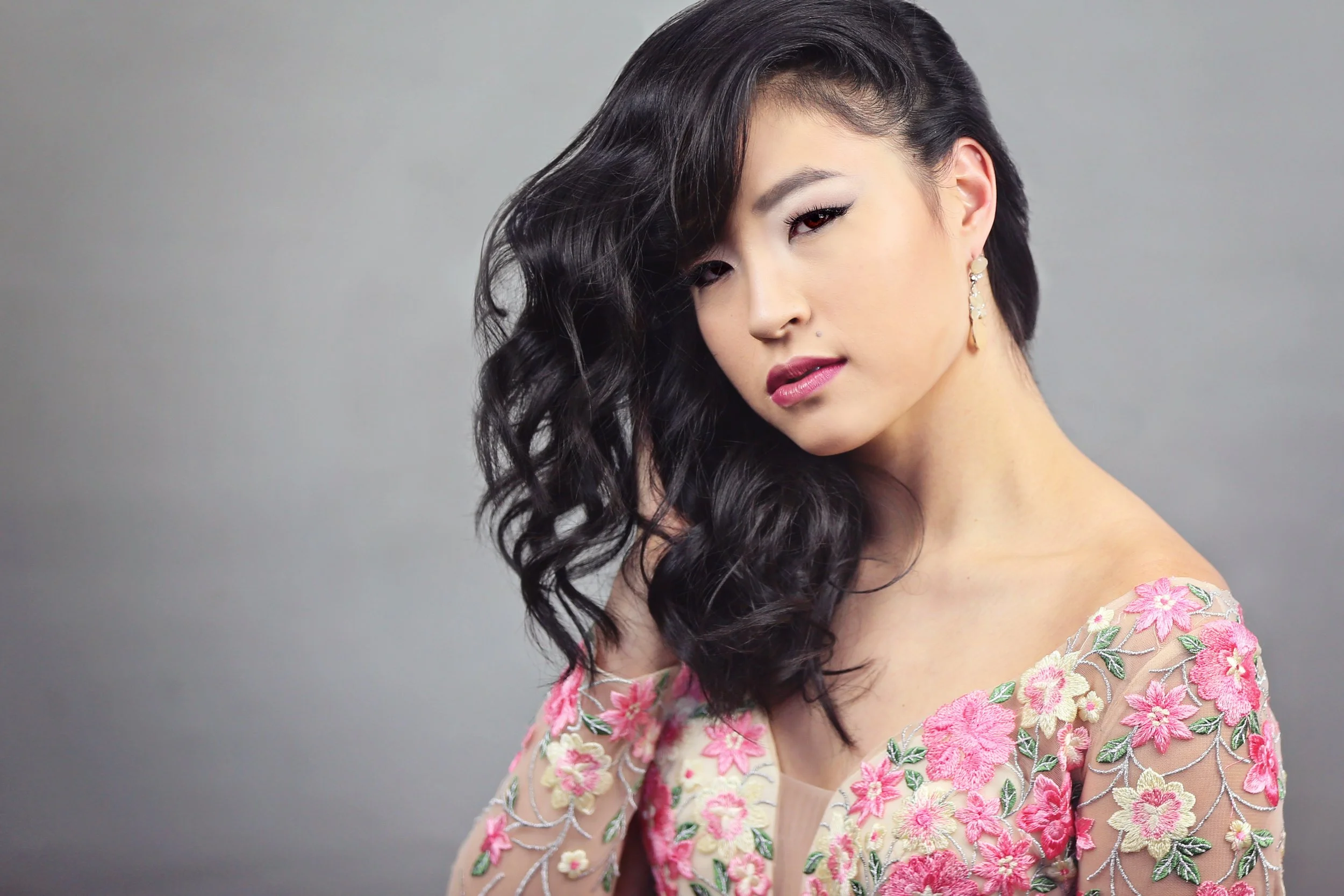 A young woman with wavy black hair, wearing a sheer dress with pink and yellow floral embroidery, and earrings, poses against a gray backdrop.