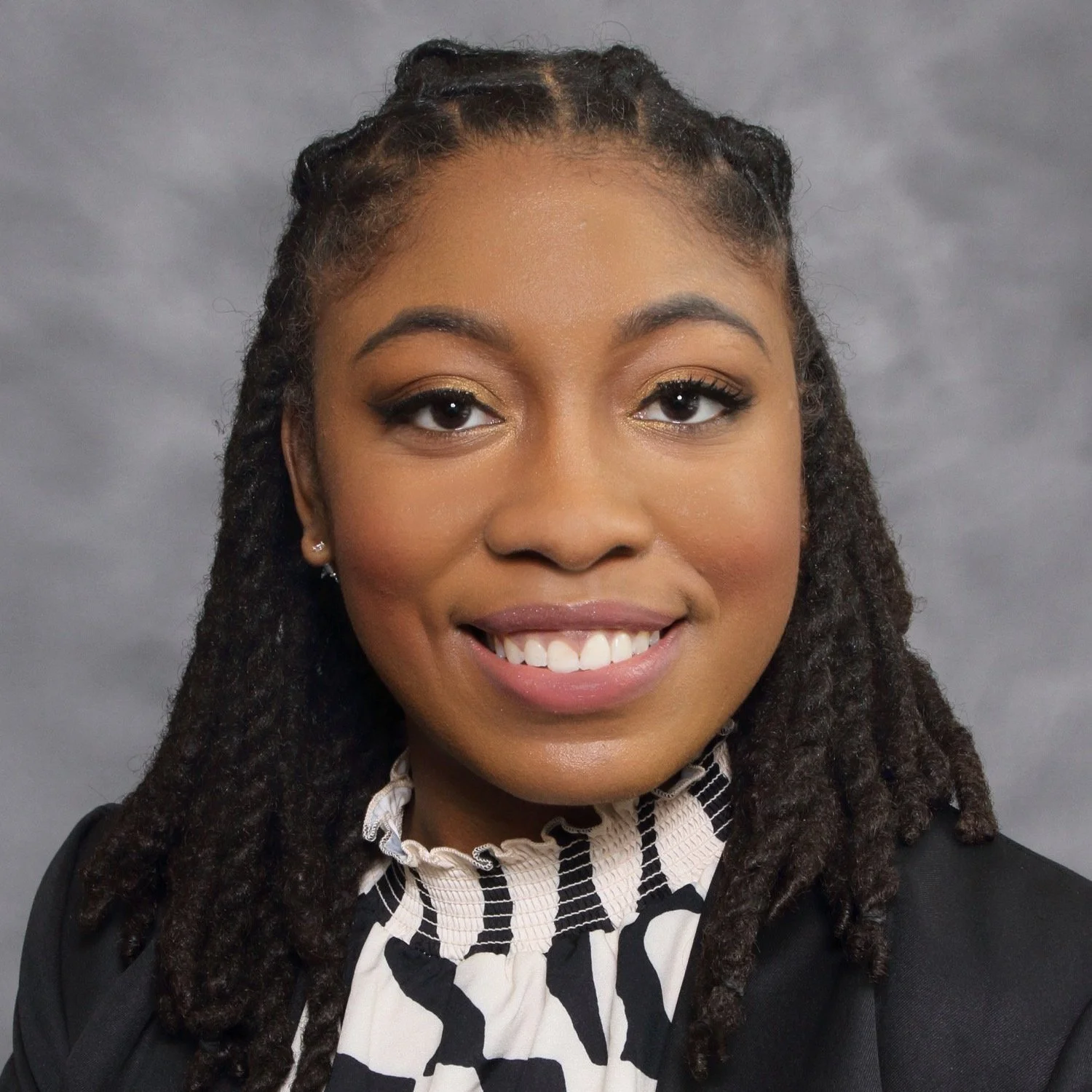 Portrait of a woman smiling with natural hair styled in dreadlocks, wearing a black blazer and a black-and-white patterned blouse, against a gray background.