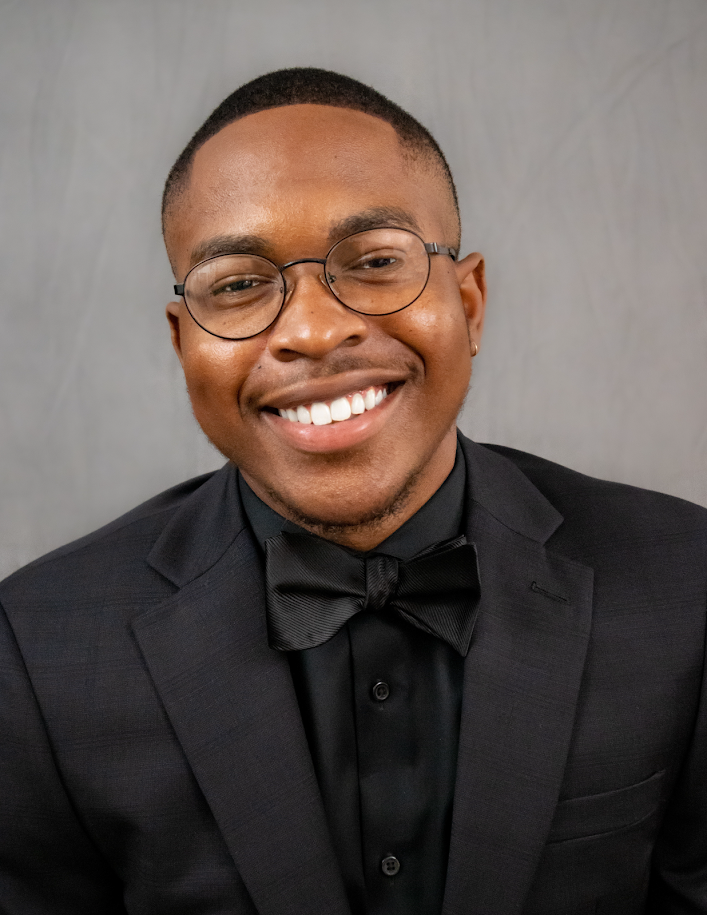 A smiling young man in a black tuxedo and bow tie, wearing glasses, against a plain light gray background.