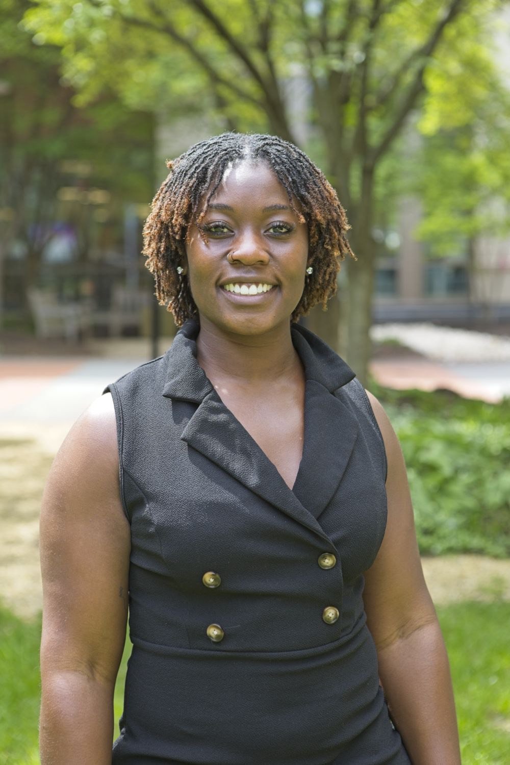 A woman with short dreadlocks smiling outdoors in front of trees and greenery.