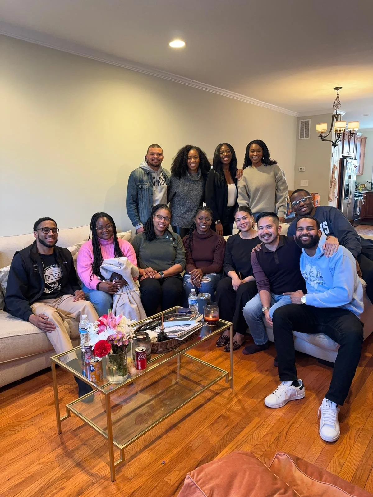 A group of fourteen young adults gathered in a living room, sitting and standing around a couch, smiling for a photo. The room has wooden floors, a glass coffee table with flowers, drinks, and snacks, and a kitchen in the background.