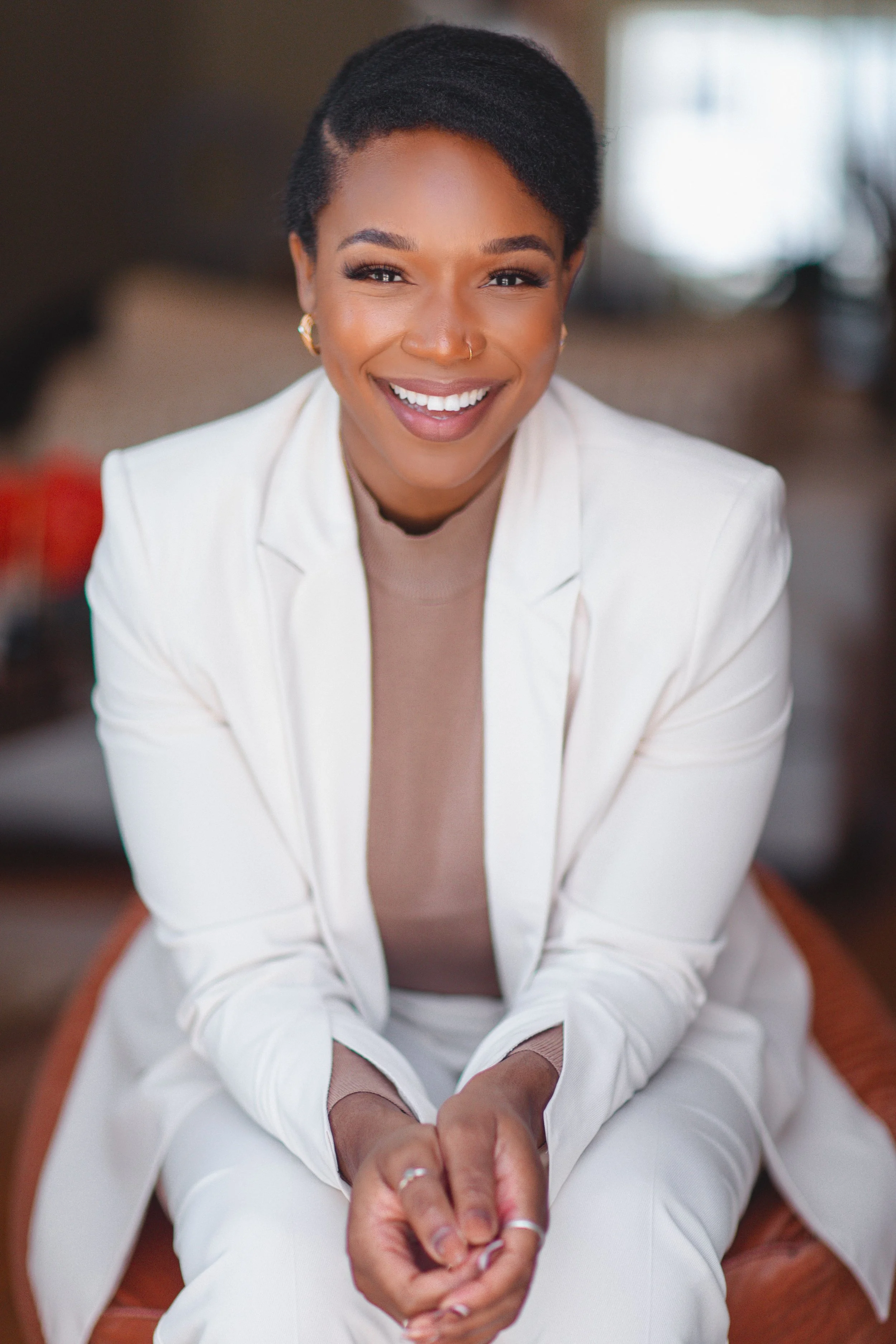 A smiling woman with short black hair, wearing a white blazer and beige top, sitting indoors.