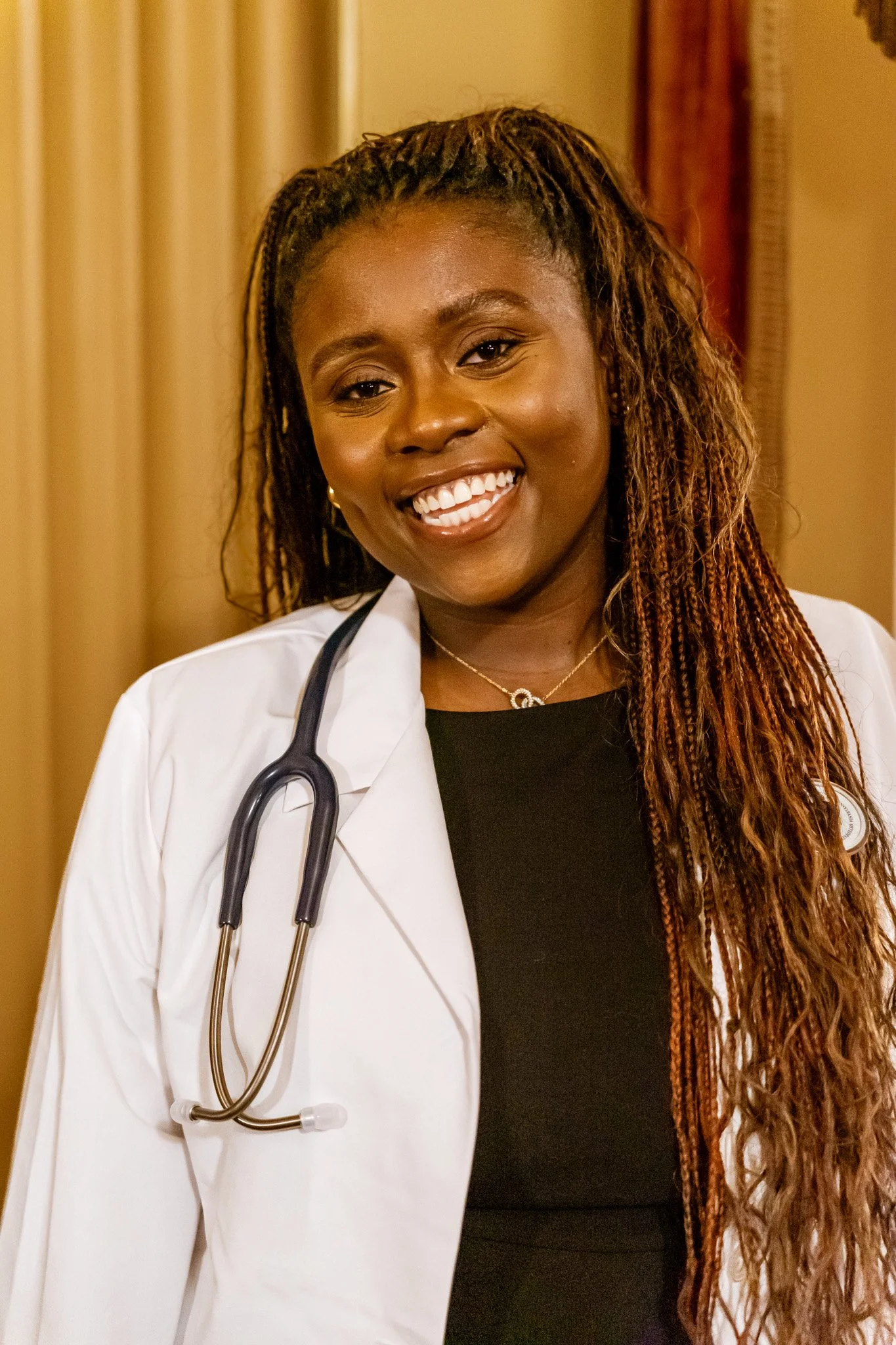 A smiling African American female doctor wearing a white coat and black shirt with a stethoscope around her neck indoors.