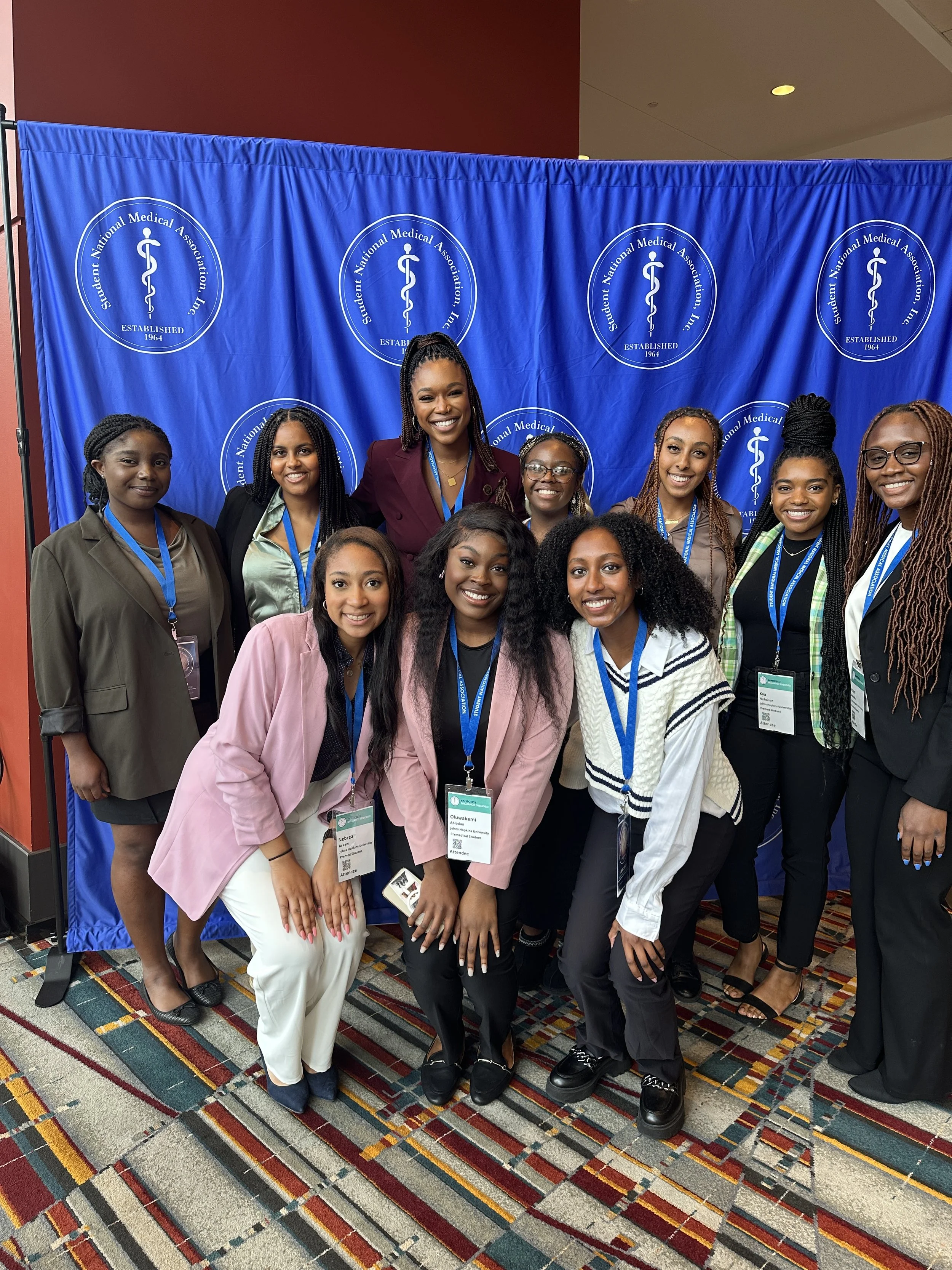 Group of nine women in professional attire posing in front of a blue backdrop with the Student National Medical Association logo at a conference.