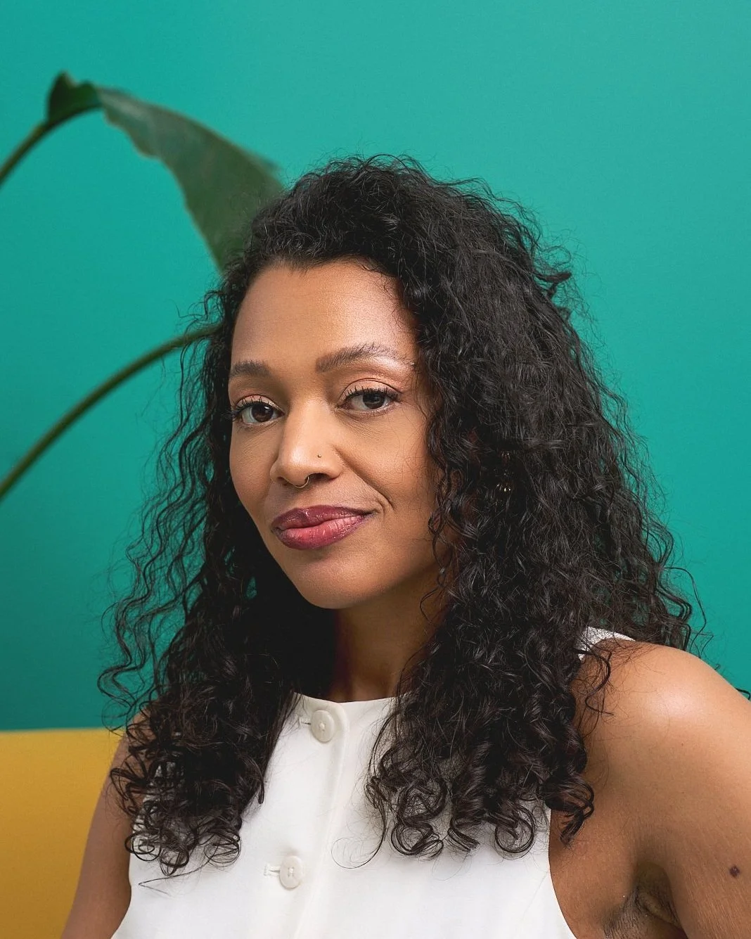 Portrait of a woman with curly black hair wearing a white sleeveless top, standing against a colorful background with green and yellow sections, near a large green leaf.