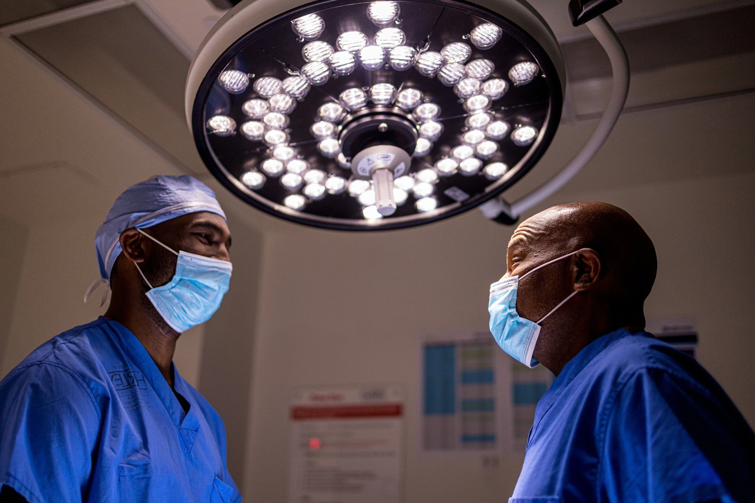 Two healthcare professionals in surgical masks and scrubs standing under bright surgical lights in an operating room.