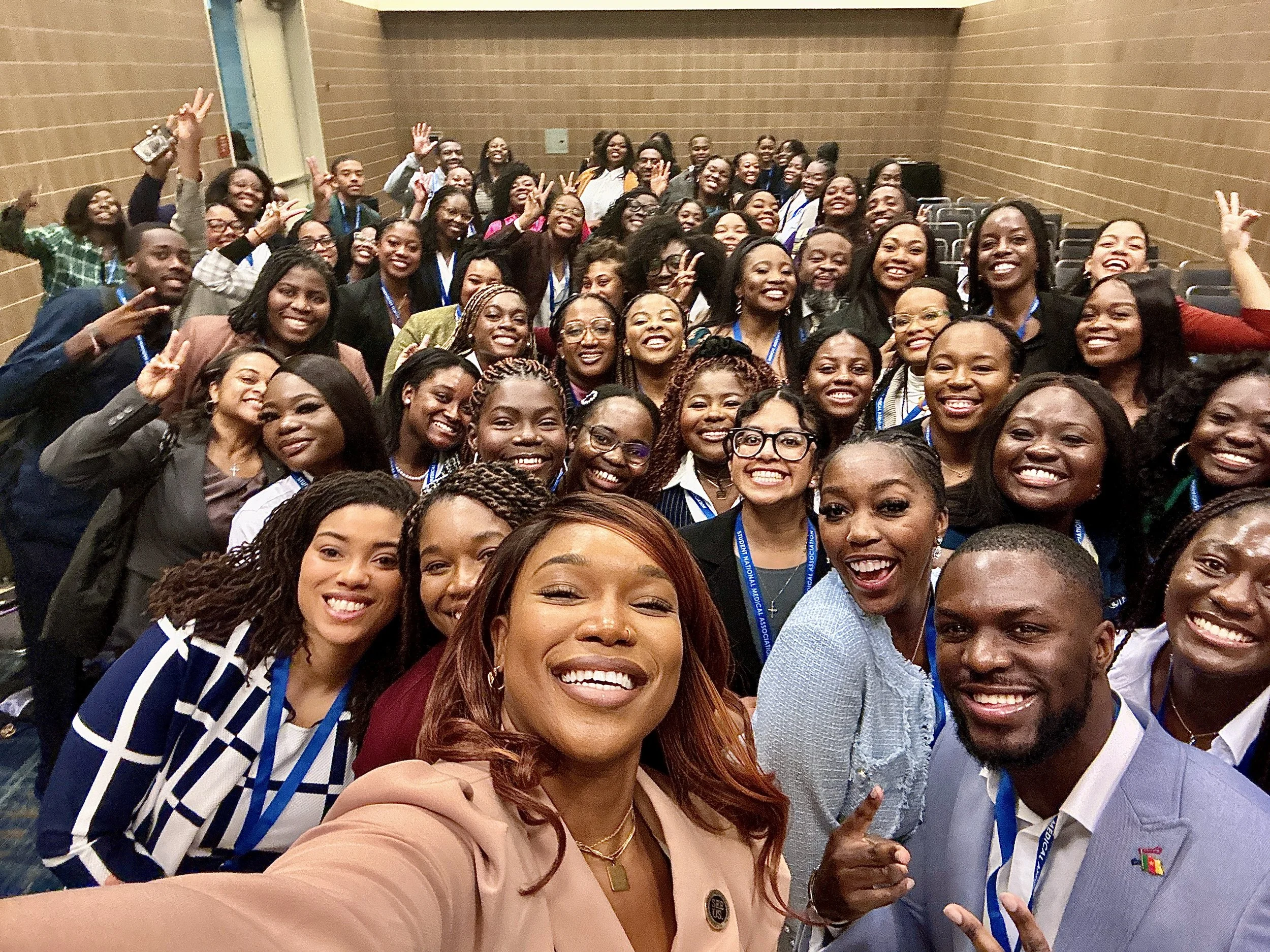 Large group of smiling people taking a selfie in a conference room with brown brick walls. The group includes men and women of diverse ages and backgrounds, many wearing conference badges.