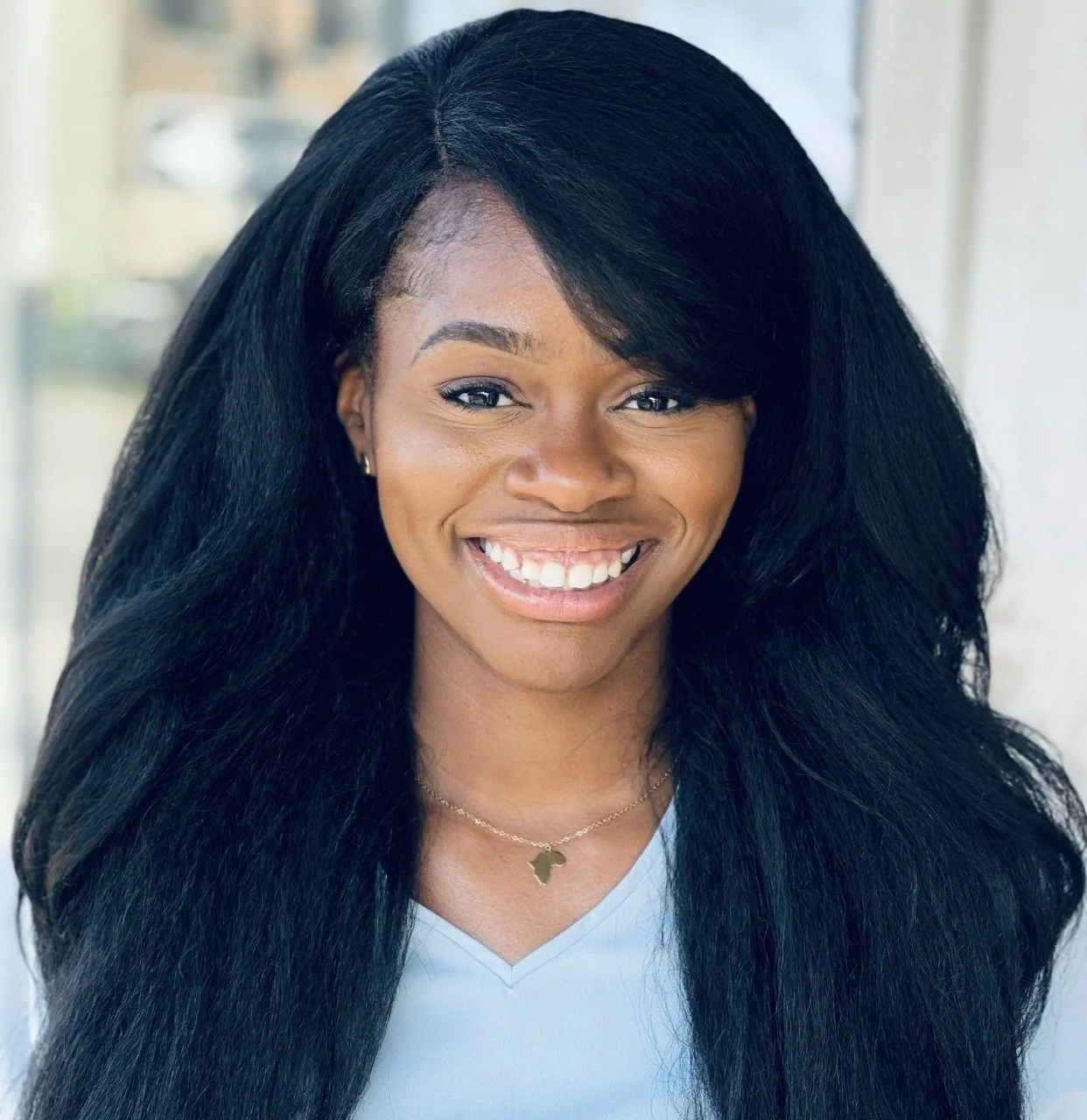 Smiling African American woman with long black hair, wearing a light blue top and a gold necklace shaped like a map of Africa.