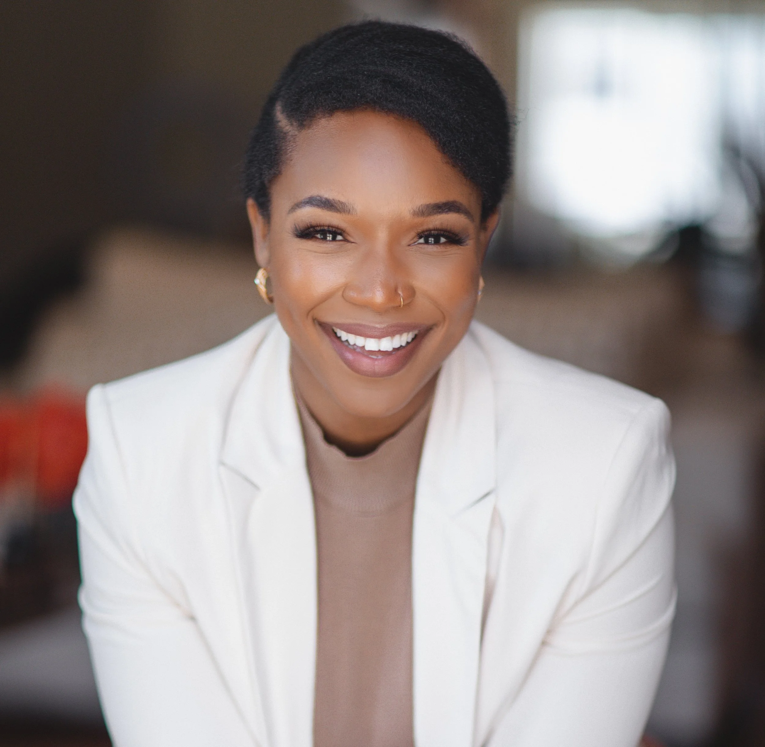 A woman with short black hair, gold hoop earrings, a nose ring, and a wide smile, wearing a beige top and a white blazer, indoors with blurred background.