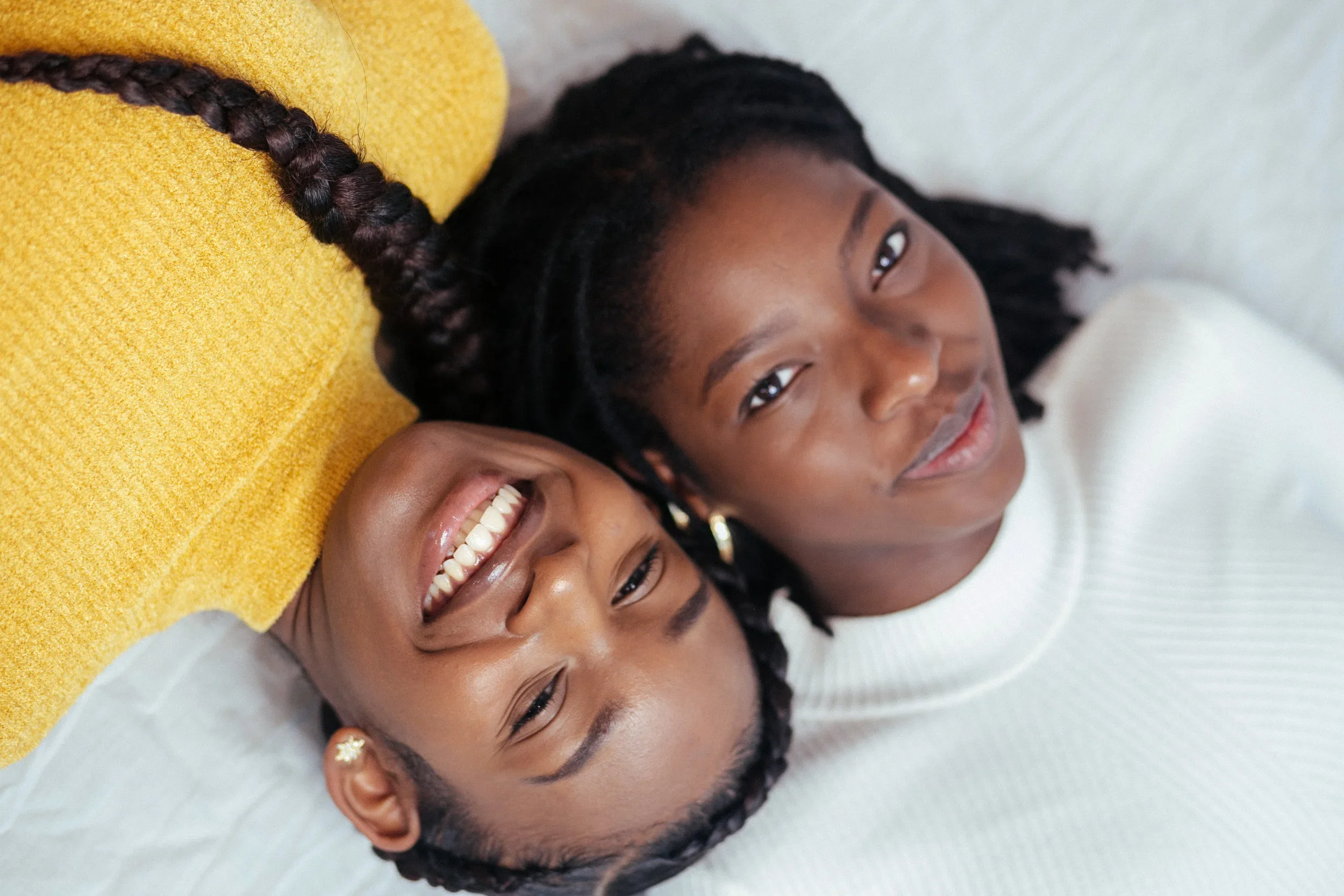 Two women lying on a white surface, one smiling and the other looking at the camera, with a yellow pillow nearby.