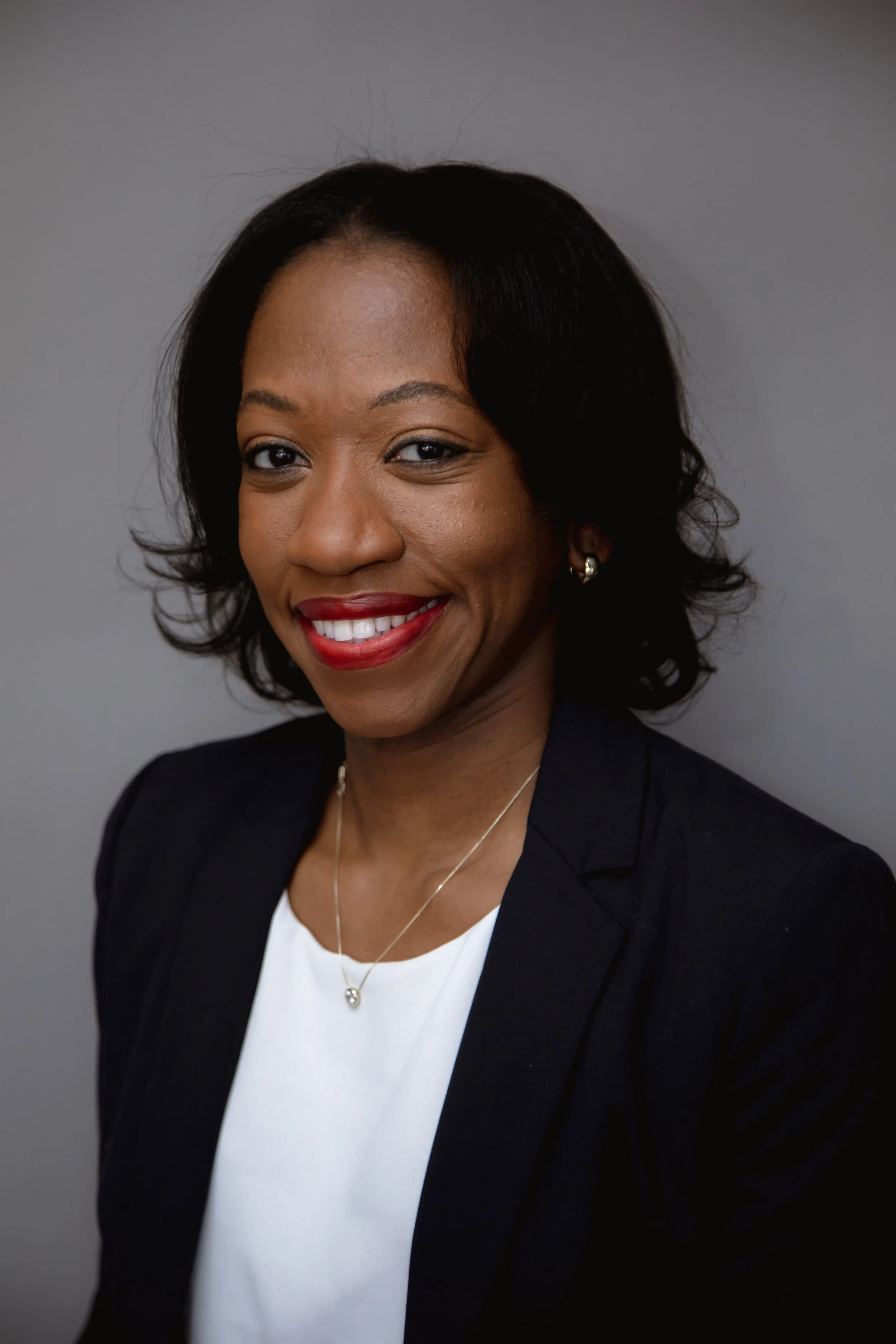 A professional woman smiling, wearing a black blazer, white top, gold jewelry, and red lipstick, against a gray background.