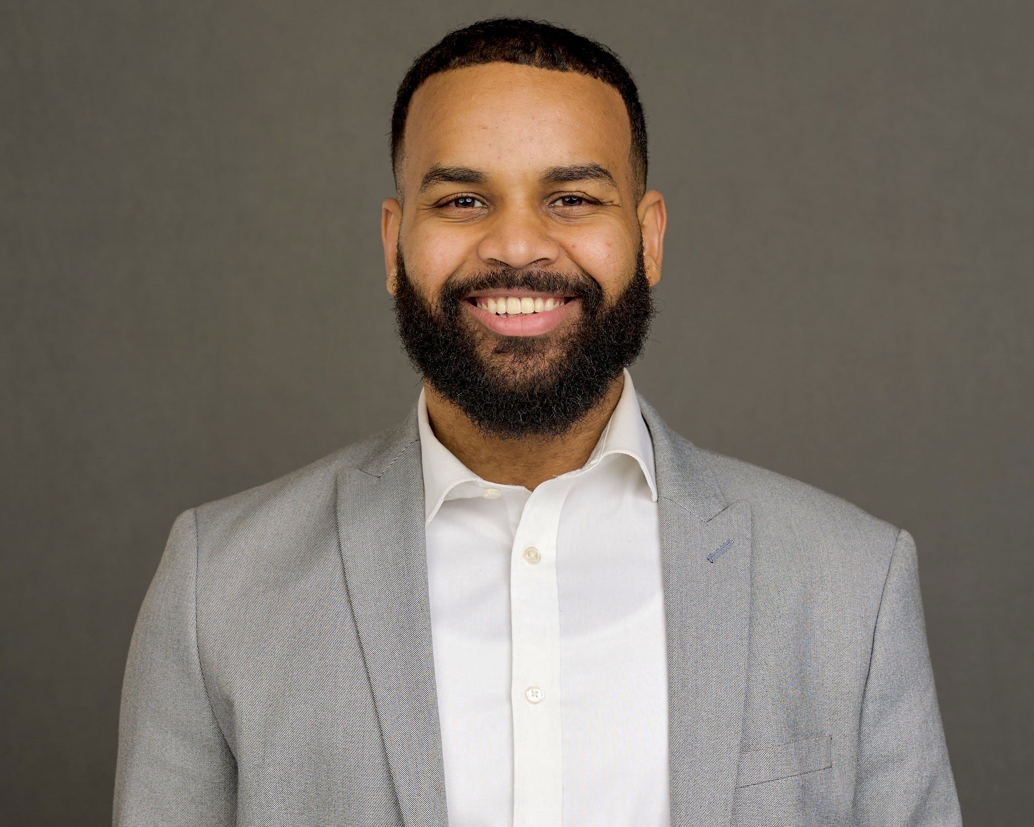 A smiling man in a light gray suit and white shirt standing against a plain gray background.