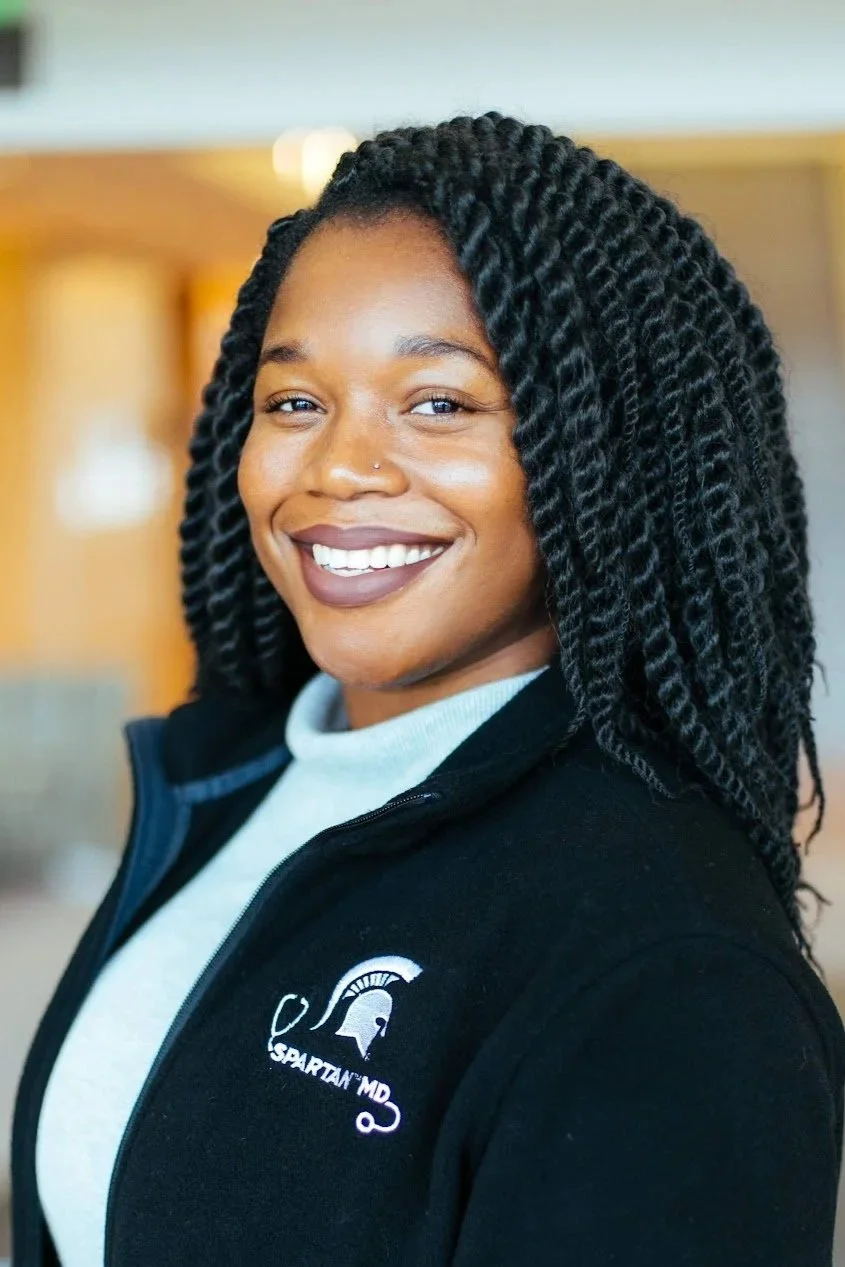 A smiling African American woman with twisted hair, wearing a black zip-up jacket with the Spartan MD logo, standing indoors.