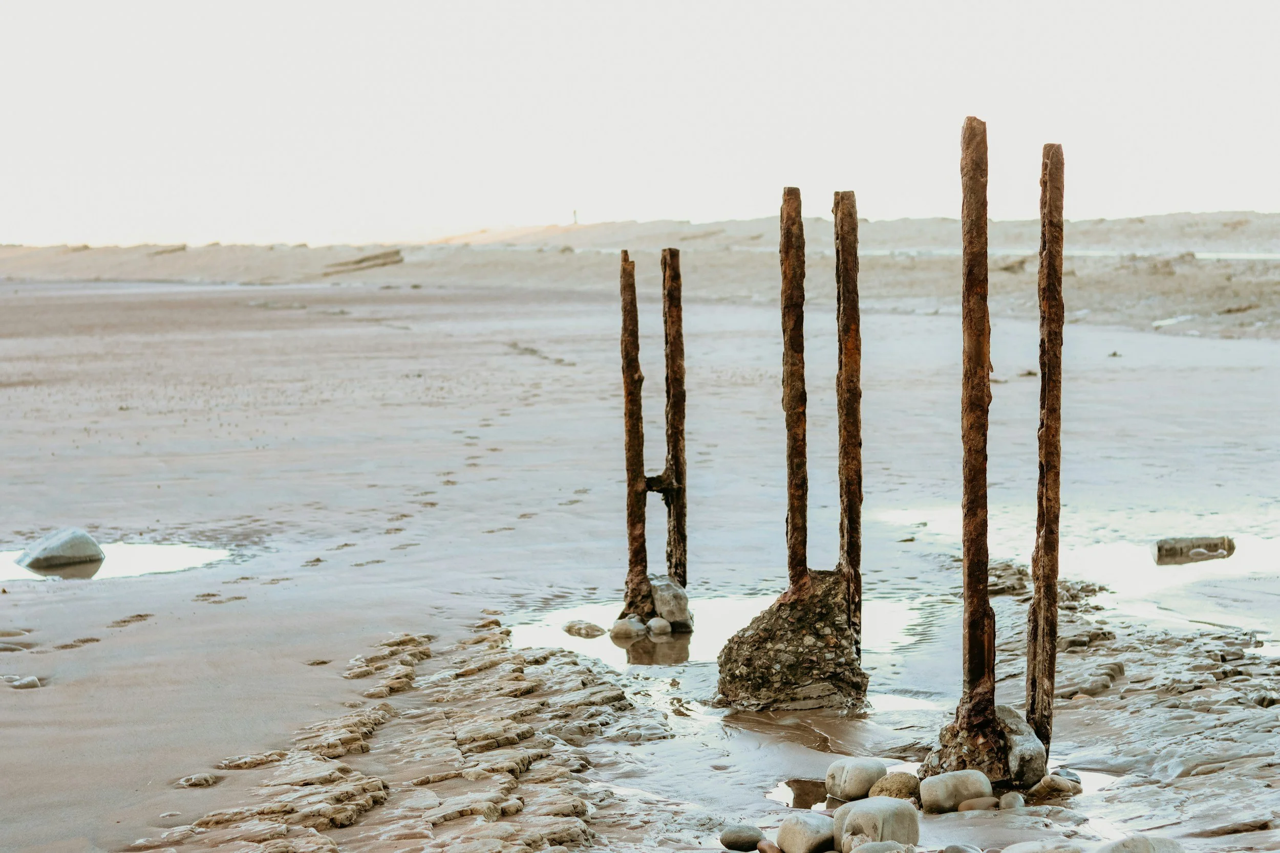 Rusty metal posts standing in shallow water along a rocky, sandy shoreline