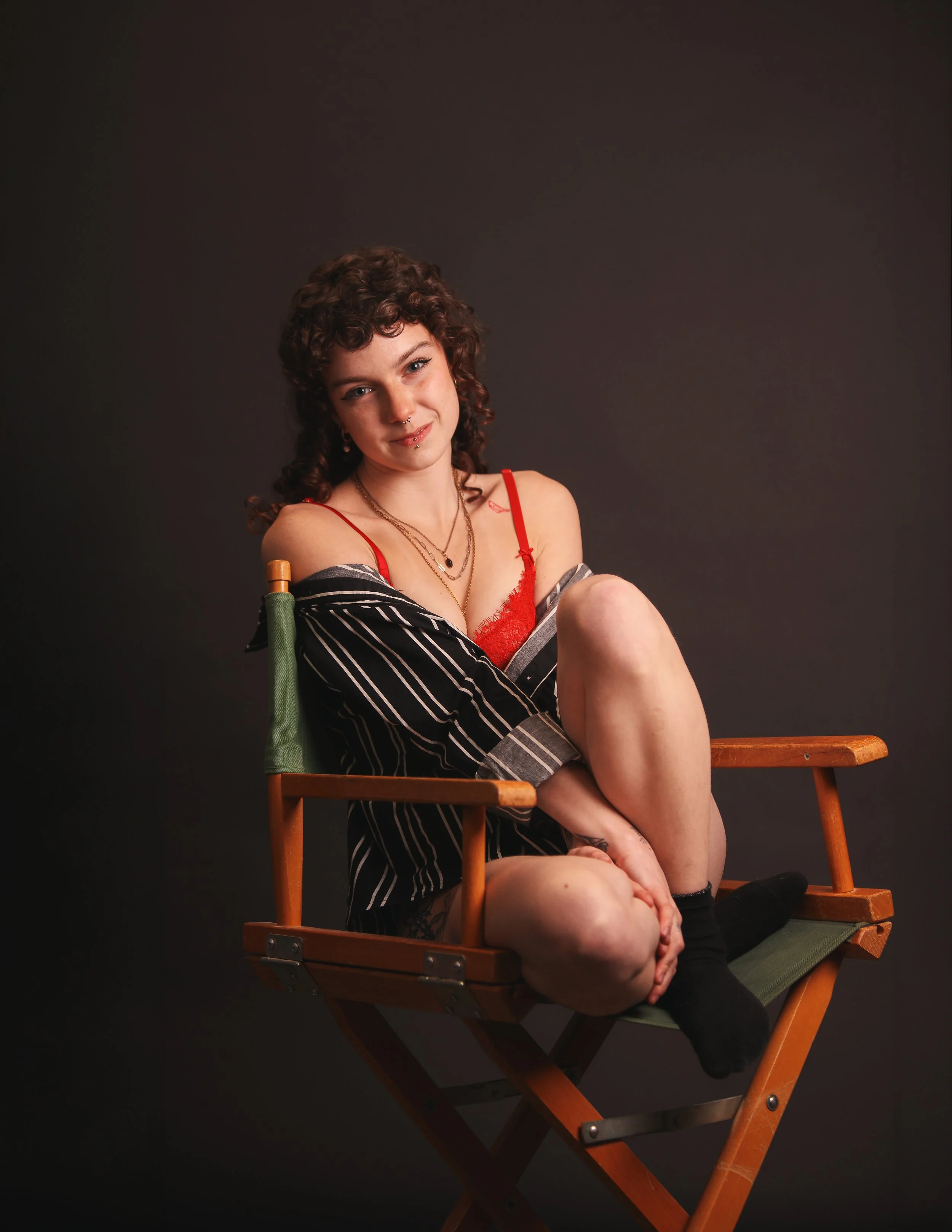 A young woman with curly brown hair and tattoos sits on a wooden director's chair against a dark background. She is wearing a red lace camisole and a black and white striped shirt off her shoulders, smiling softly at the camera.