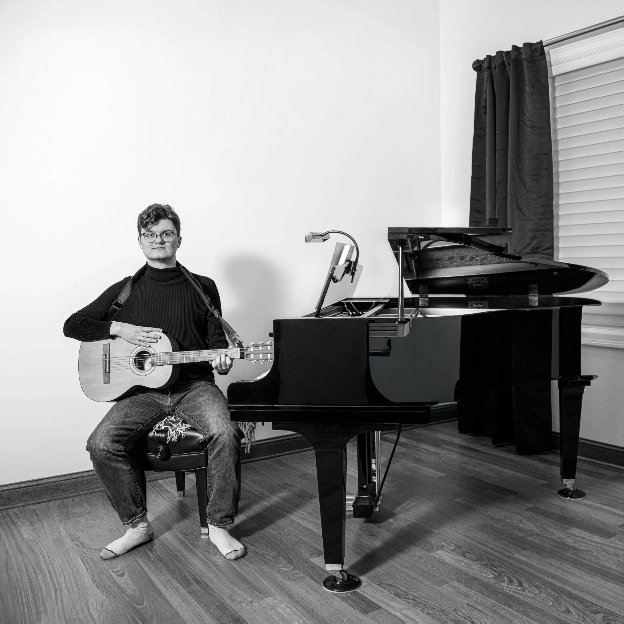 A person with short hair, glasses, and a black turtleneck sitting on a bench, holding a guitar, in front of a grand piano in a room with wooden flooring and a white wall.