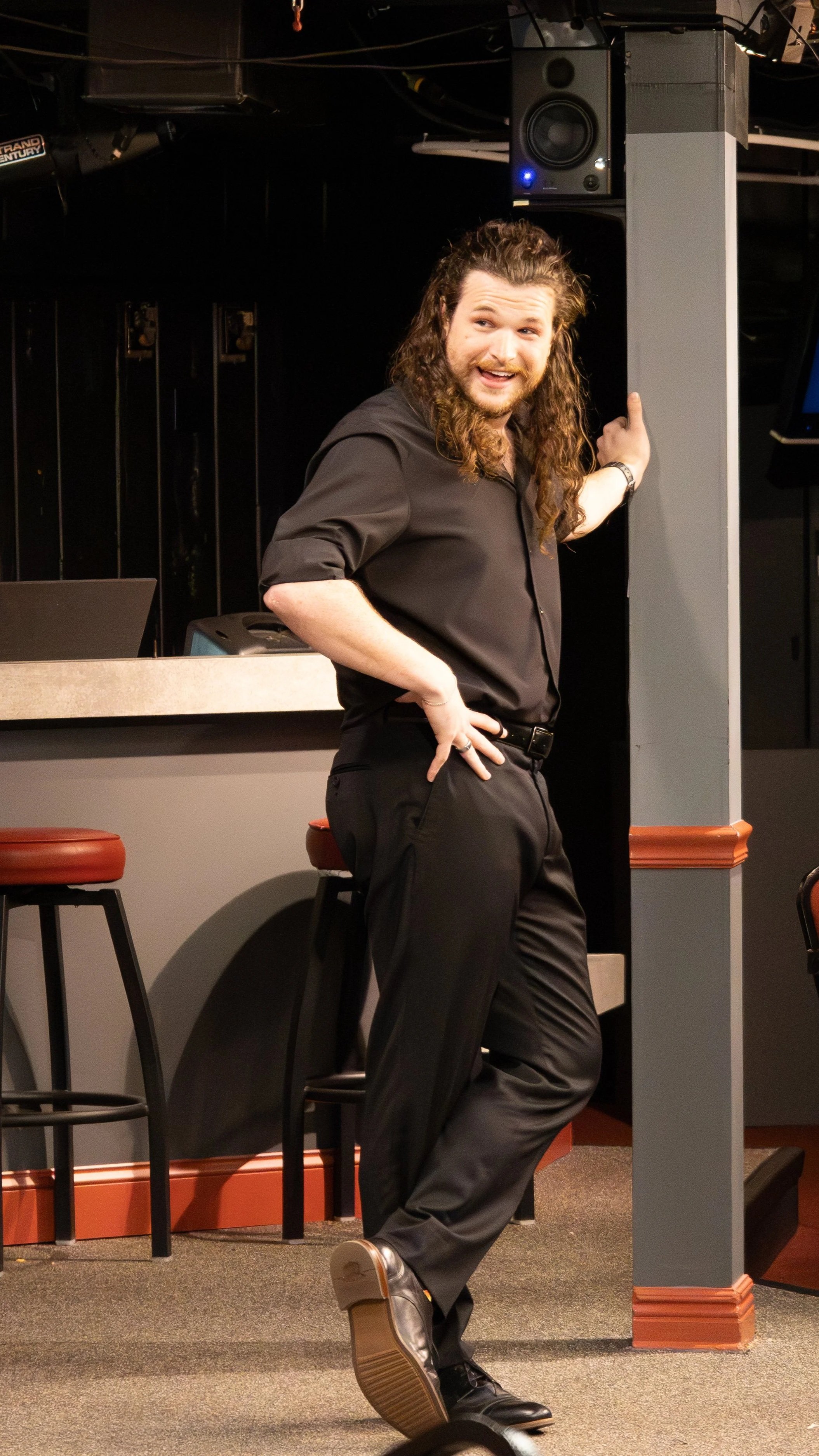 A man with long curly hair, beard, and black clothing stands in a bar or club, leaning against a bar with one hand on a speaker and smiling, with stools and a counter behind him.