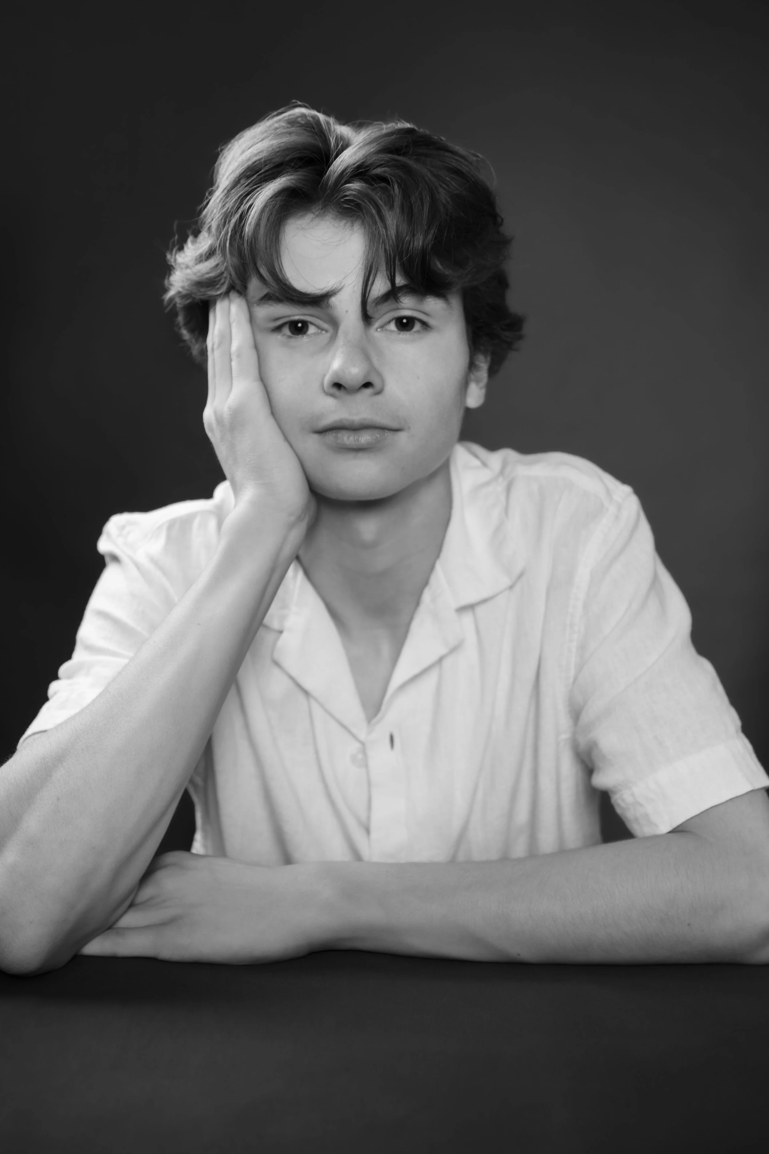 Black and white portrait of a young man with wavy hair, resting his head on his left hand, and staring at the camera with a neutral expression.