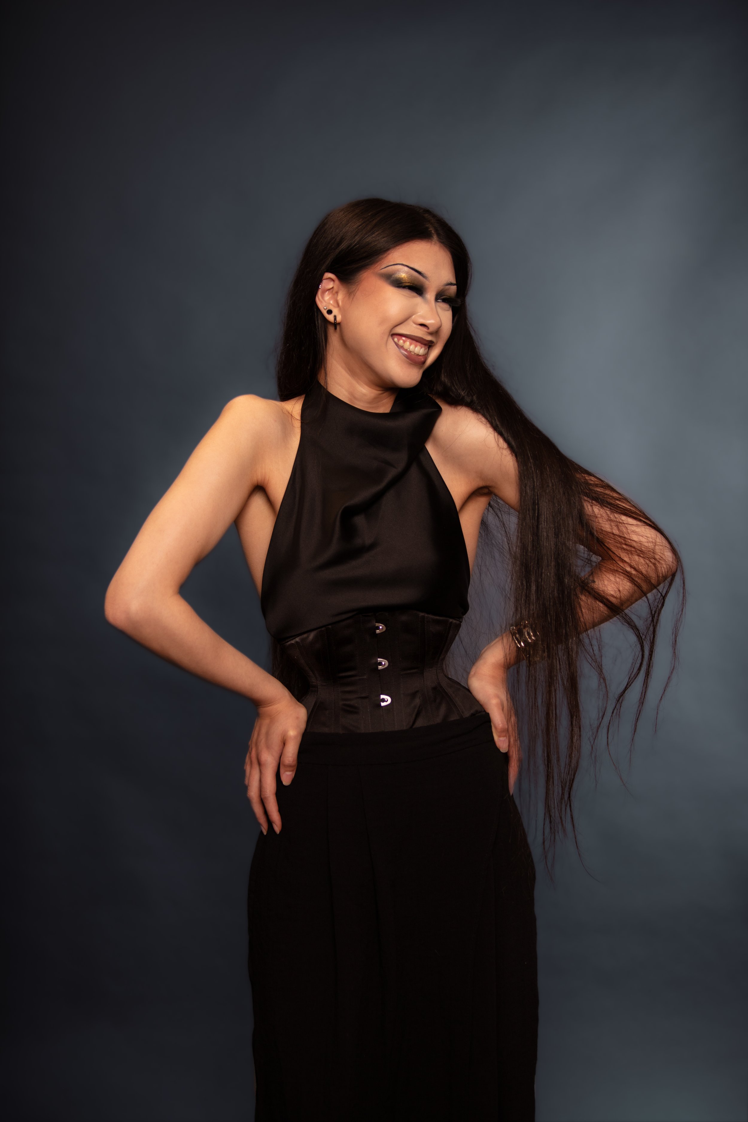 A woman with long dark hair, makeup, and earrings, wearing a black sleeveless top and skirt, smiling with her eyes closed, standing against a dark background.