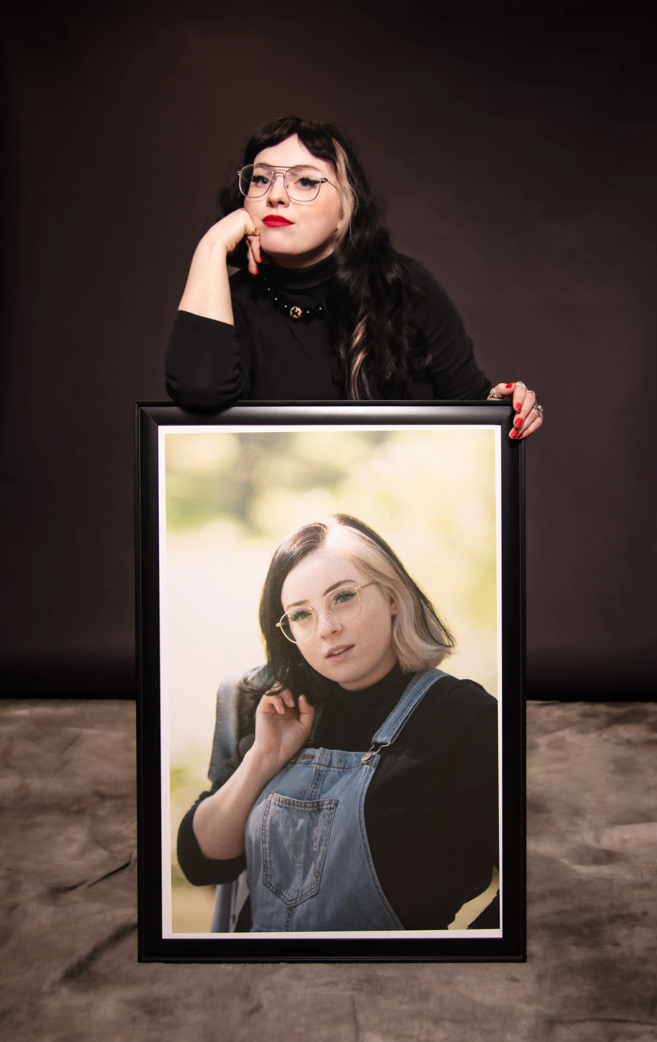 A woman with long black hair, glasses, and red lipstick sits at a table, resting her chin on her hand, with a framed photo of another woman in front of her. The background is dark and plain.
