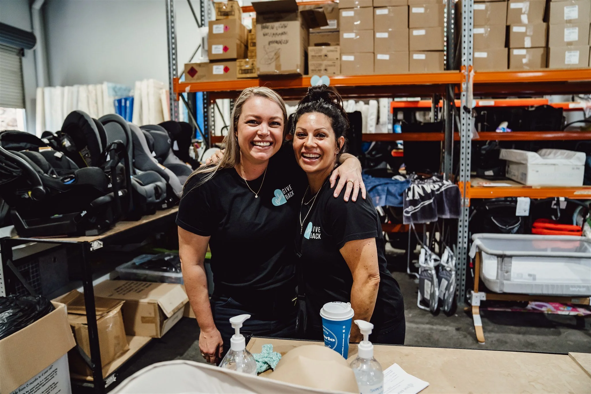 Two volunteers in the baby give back warehouse in front of donated items
