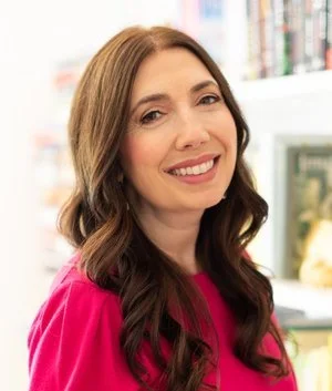 Smiling woman with long brown hair wearing a pink top, standing indoors.