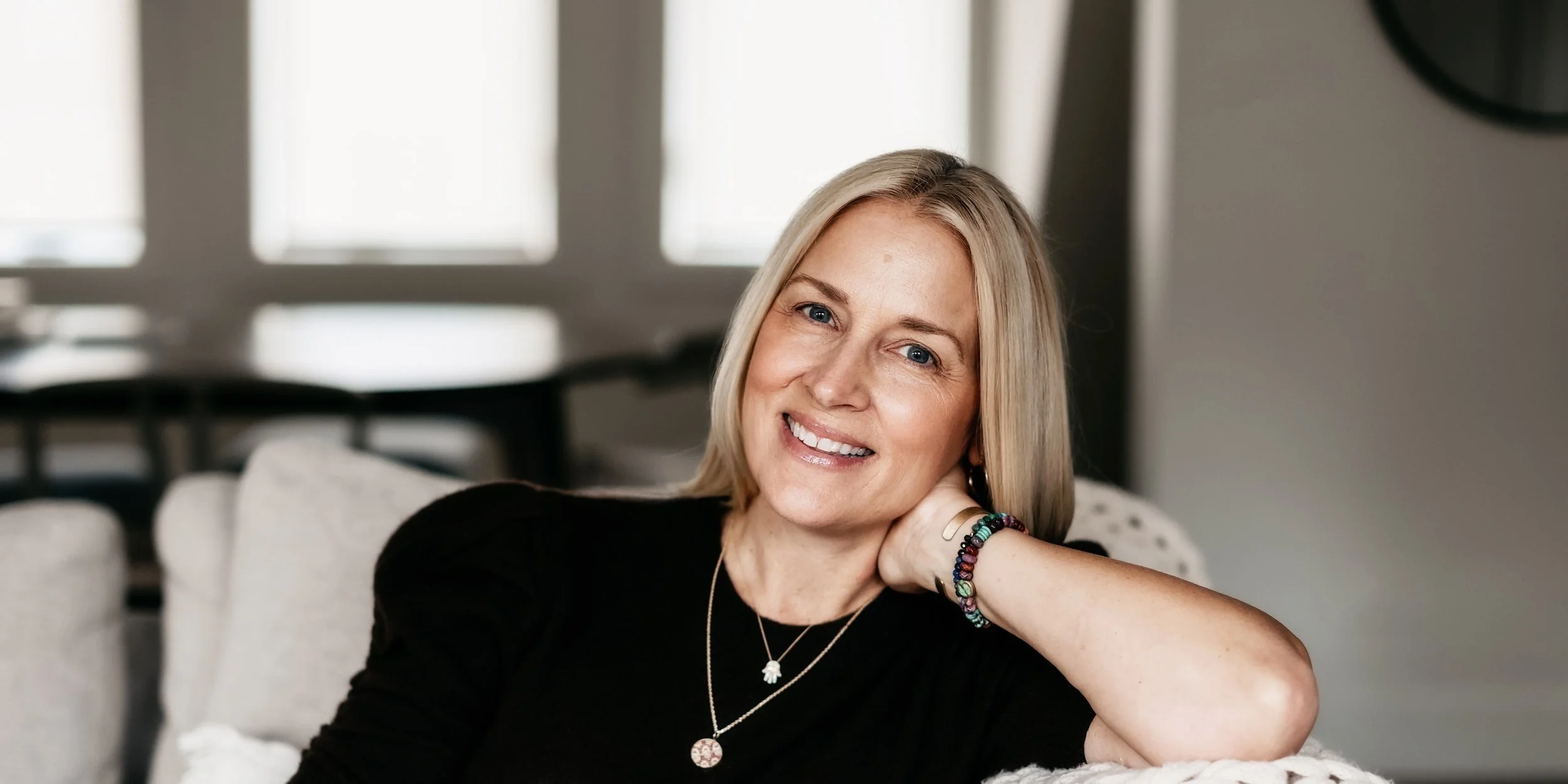 Smiling woman with blonde hair leaning on a couch, wearing a black top, necklace, and bracelets.