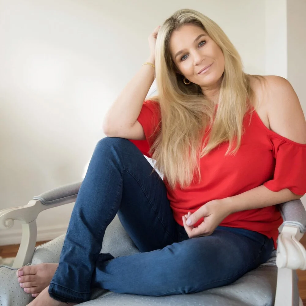 A woman with long blonde hair sitting comfortably on a chair, wearing a red off-the-shoulder top and blue jeans.