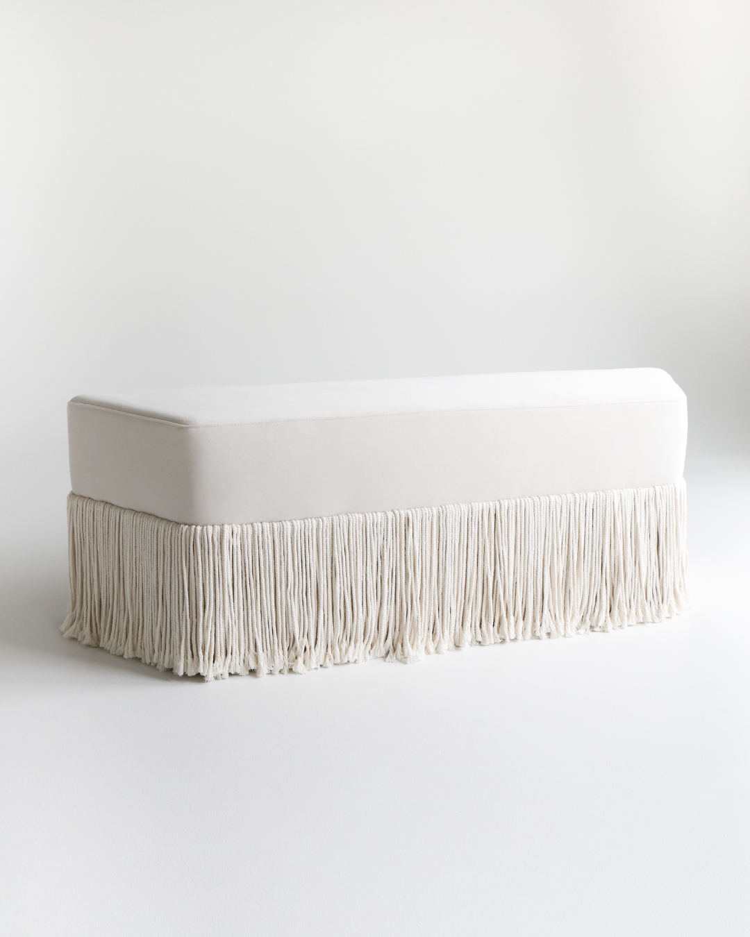 A white upholstered bench with a fringe skirt, placed against a white background.