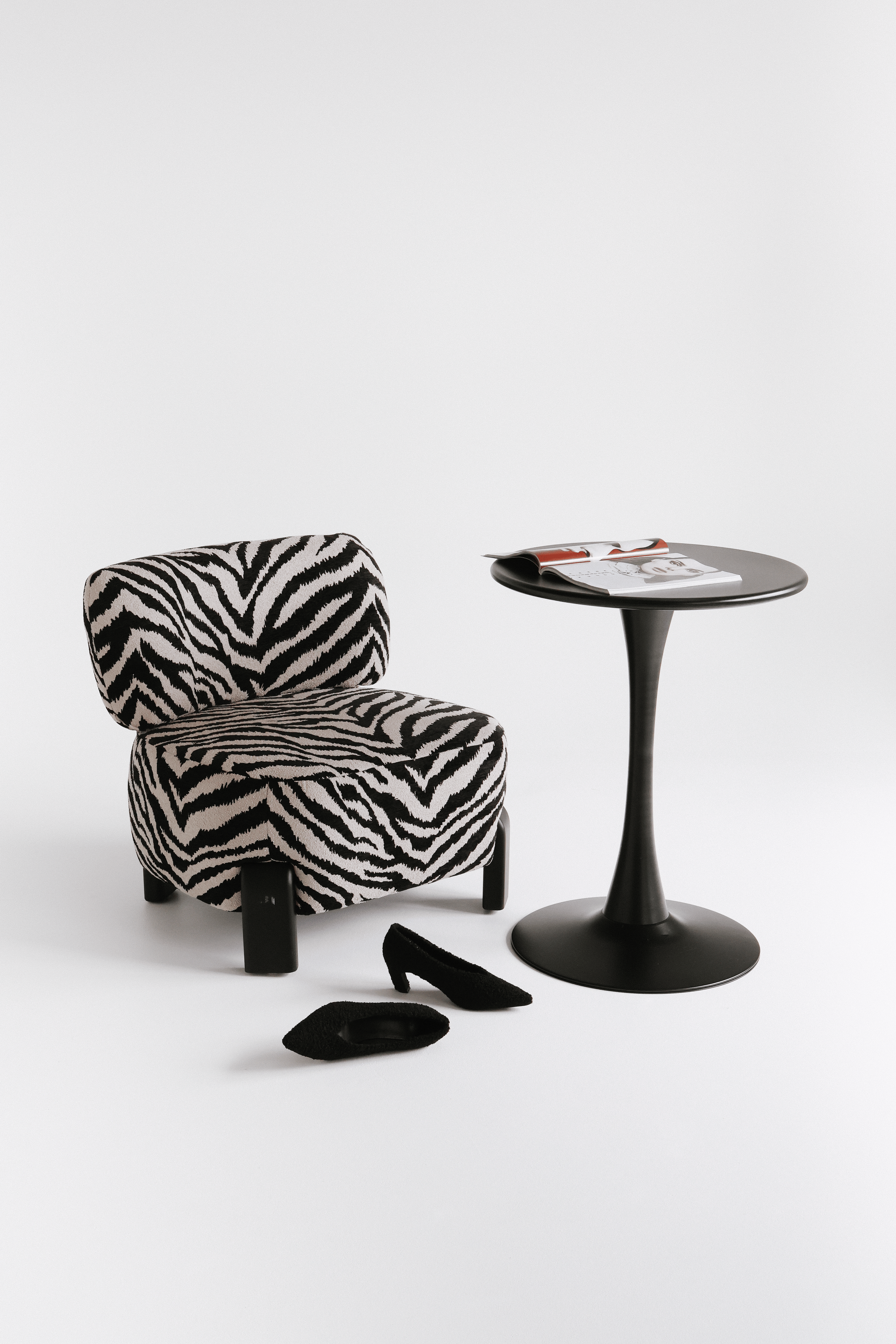 A black and white zebra print chair, black high heel shoes, a magazine on a black round side table, all set against a plain white background.