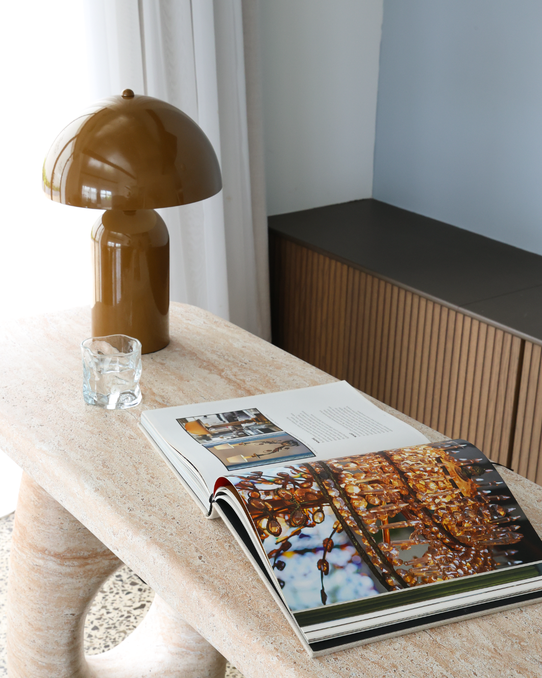 A beige stone table with an open magazine showing images of modern interior design and decor. A brown modern table lamp, a glass of water, and a white curtain are on the left side of the table. A wooden cabinet with vertical slats is against the wall behind the table.