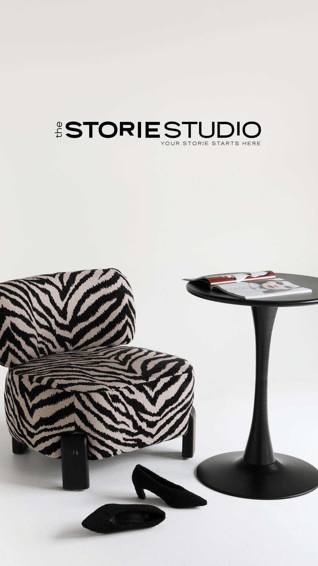 A black and white zebra-print chair next to a black round side table with magazines on it, black slippers and a black heel on a white background, with the text 'the STORY STUDIO YOUR STORY STARTS HERE' on the wall.