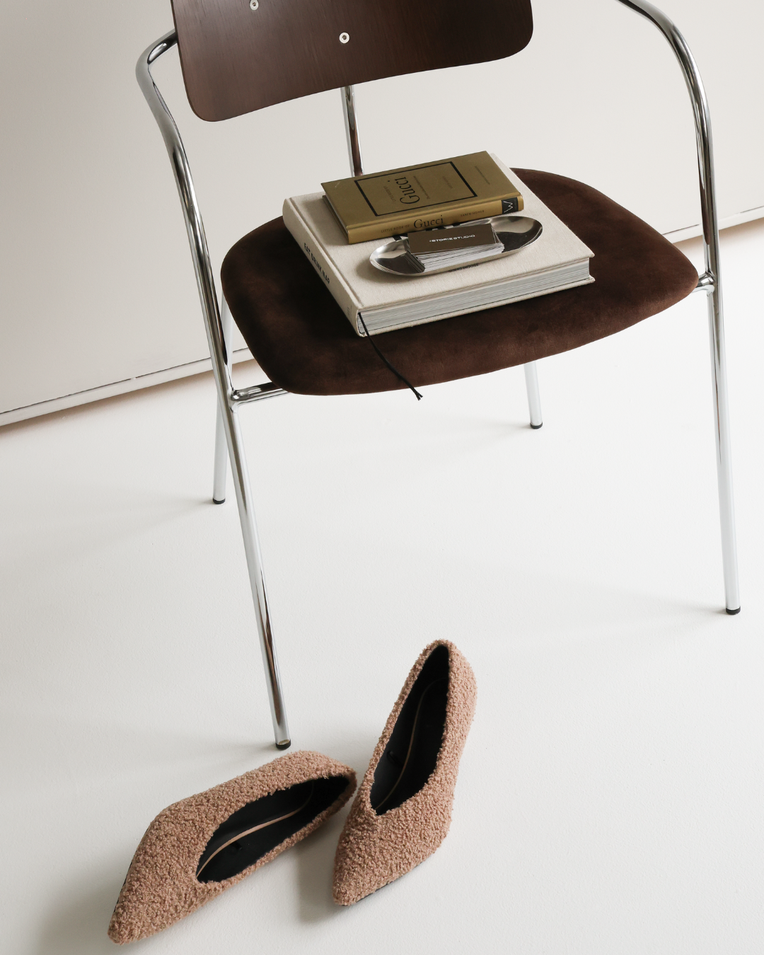 A modern chair with a dark brown seat and a wooden backrest, holding a stack of books and a pair of sunglasses. In front of the chair are a pair of pink textured high-heeled shoes placed on a white surface.
