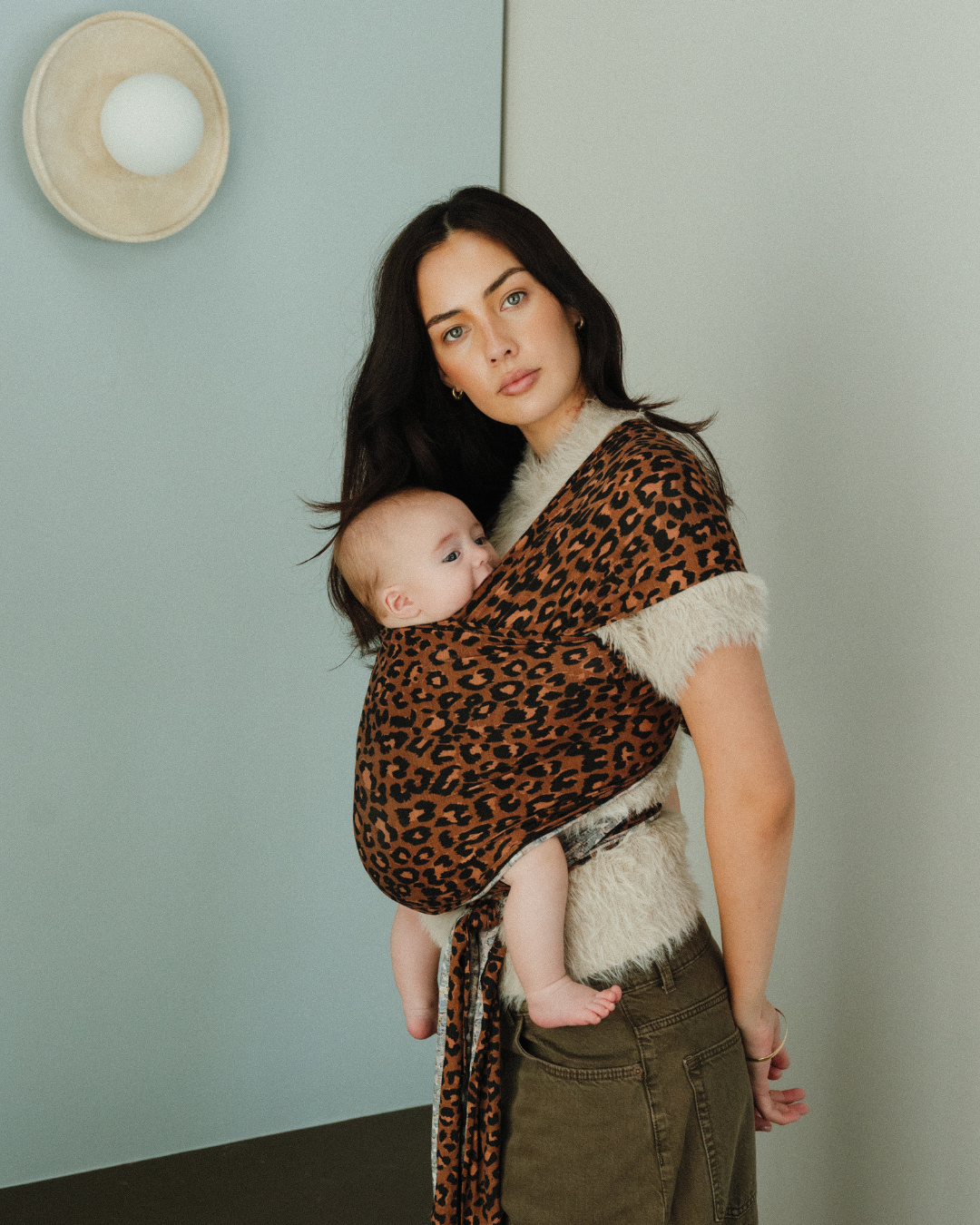 Mother and baby in a Chekoh baby carrier at the Storie Studio - background is a blue wall with wall sconce, naturally lit