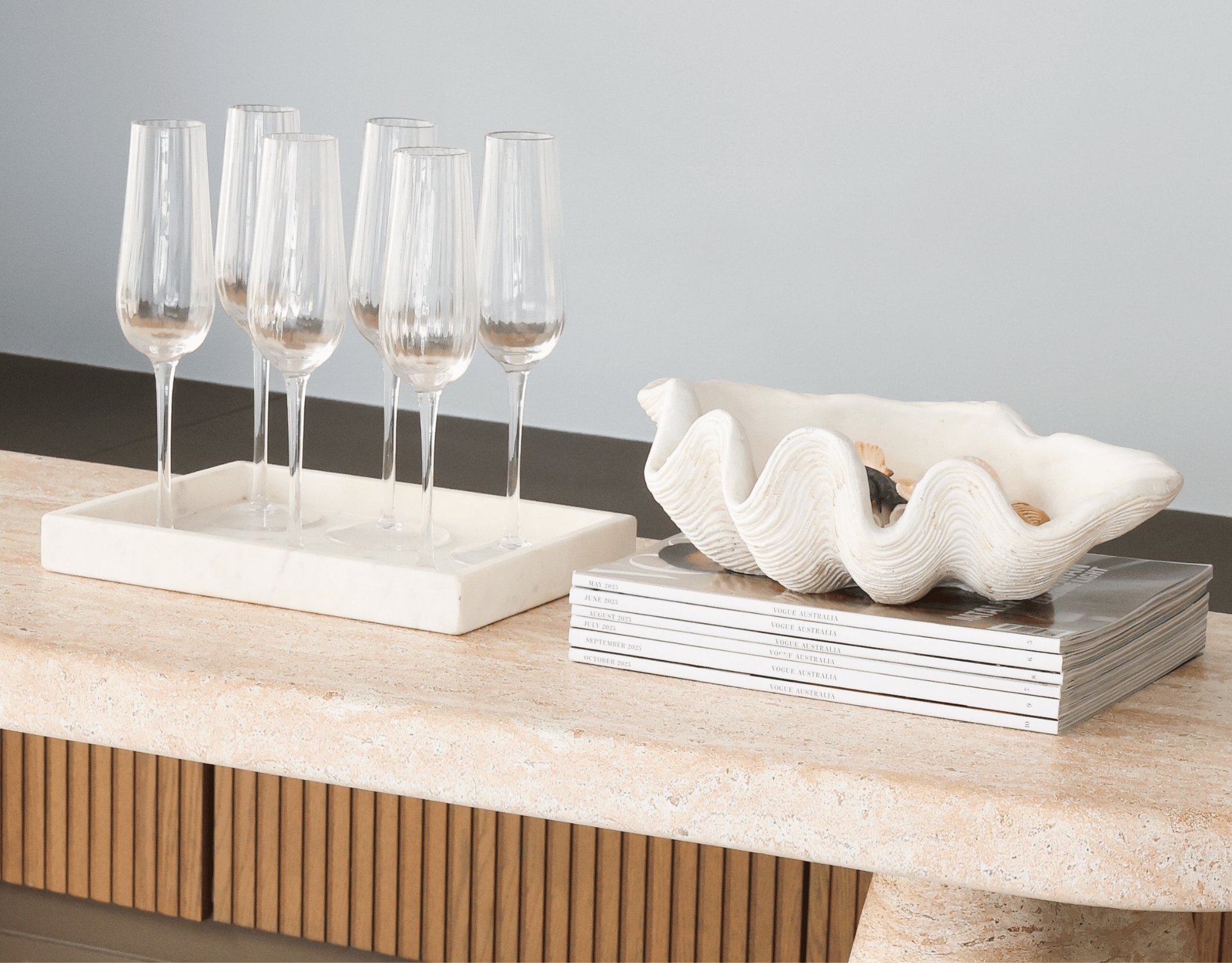 A set of six empty champagne flutes on a white marble tray, and a large shell-shaped bowl with assorted jewelry or accessories inside, placed on a stack of magazines on a wooden surface.