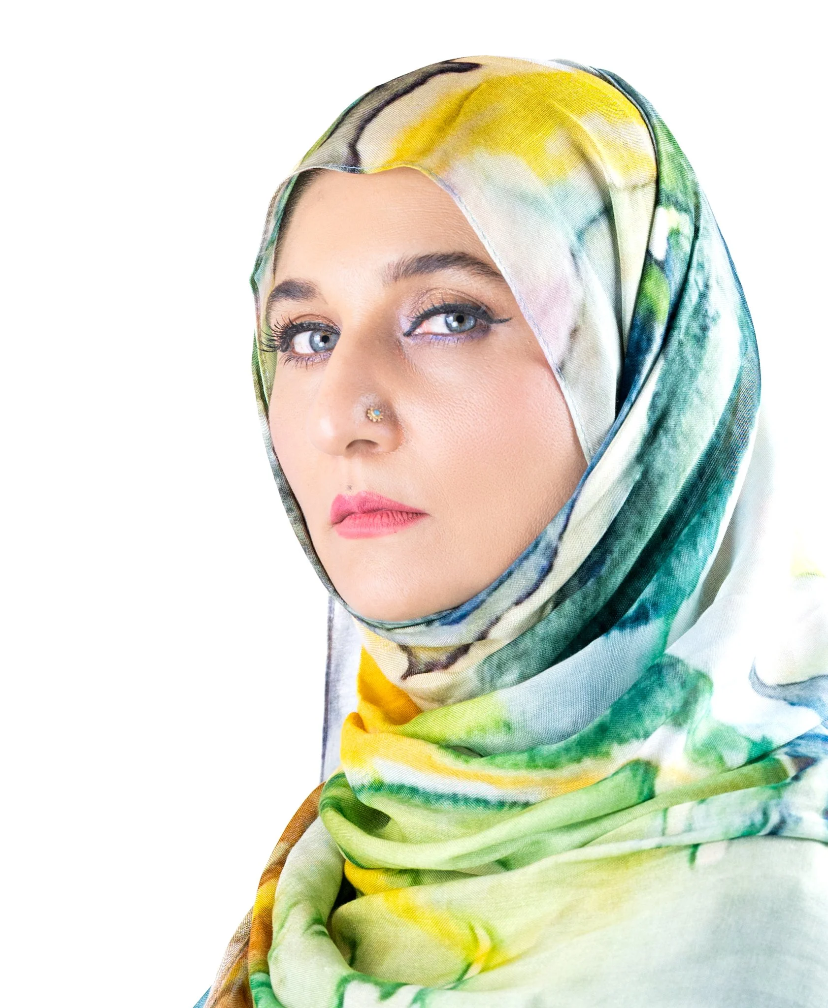 A woman with blue eyes wearing a colorful, patterned hijab and makeup, looking at the camera against a white background.