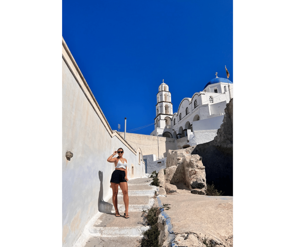 Solo female traveller sitting by a blue door beneath a traditional whitewashed church in Santorini, showcasing the peaceful, picturesque side of the island for women travelling alone.