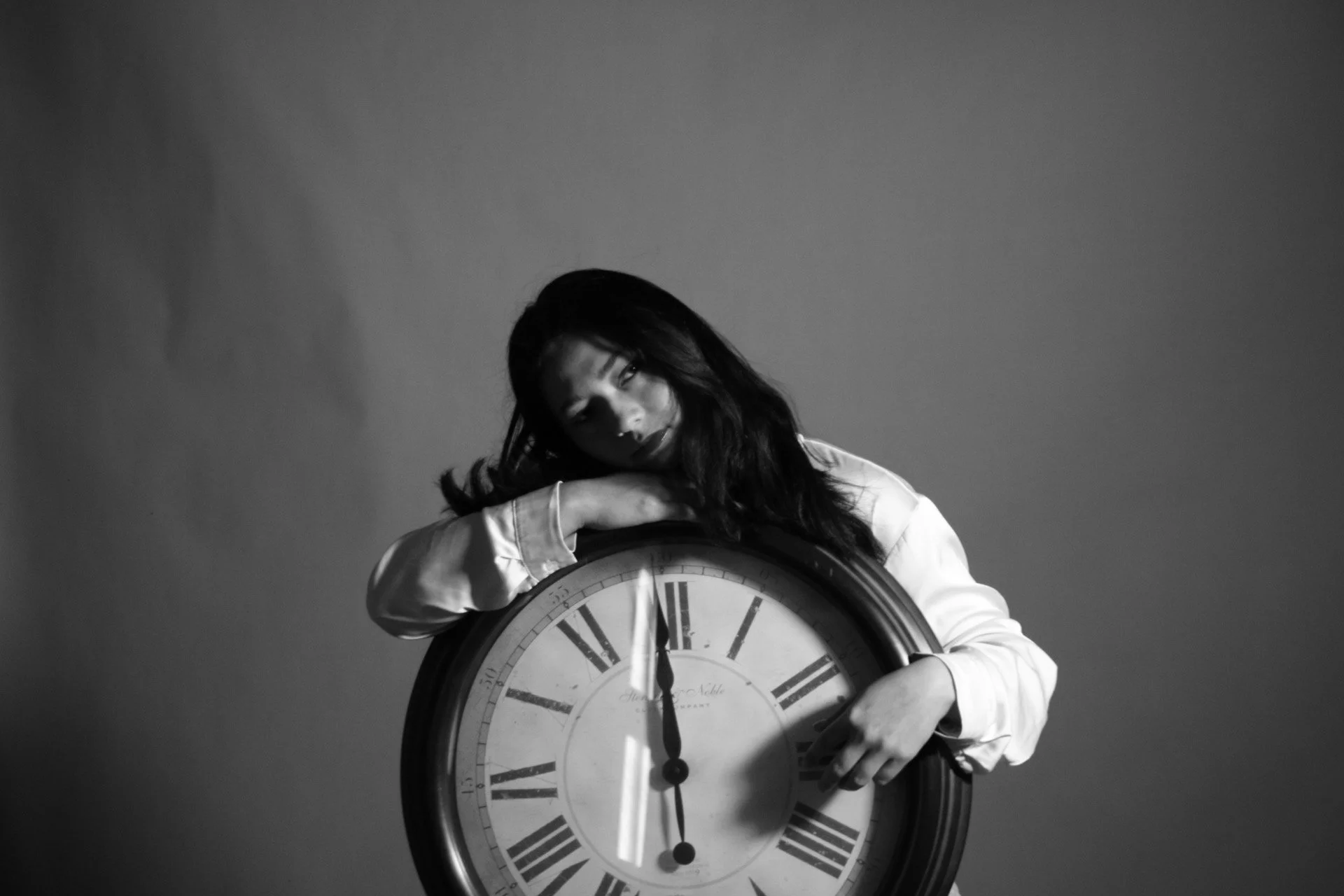Una mujer con cabello largo y oscuro, vestida con una camisa clara, apoya la cabeza en su brazo doblado sobre un gran reloj de pared con números romanos, en un fondo oscuro. La imagen es en blanco y negro.