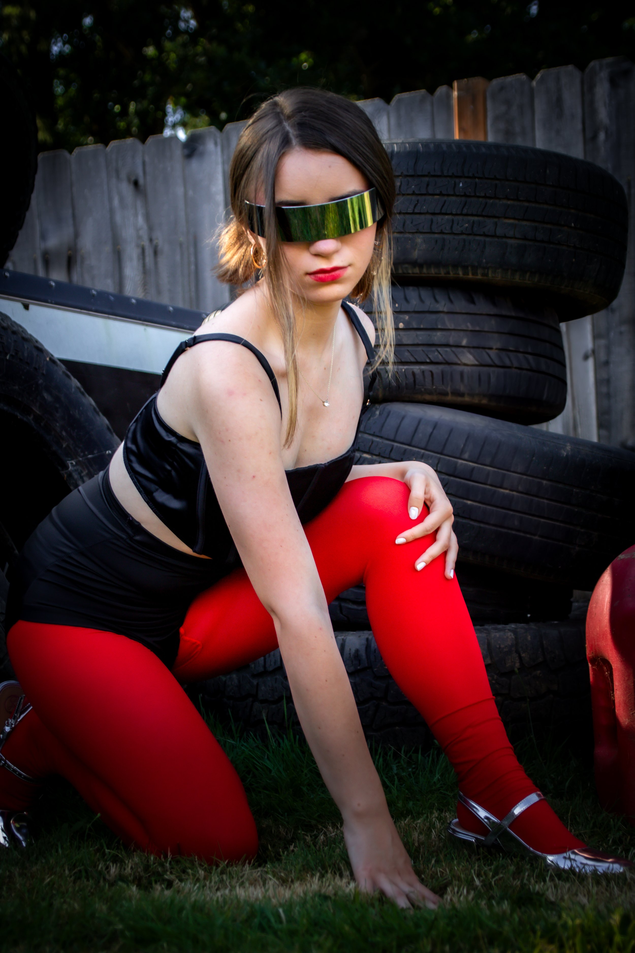 Editorial-style outdoor portrait featuring a model kneeling on grass in front of stacked tires—wearing red leggings, silver shoes, and a black top, set against a rustic wooden fence. The image evokes themes of contrast, strength, and expressive fashi