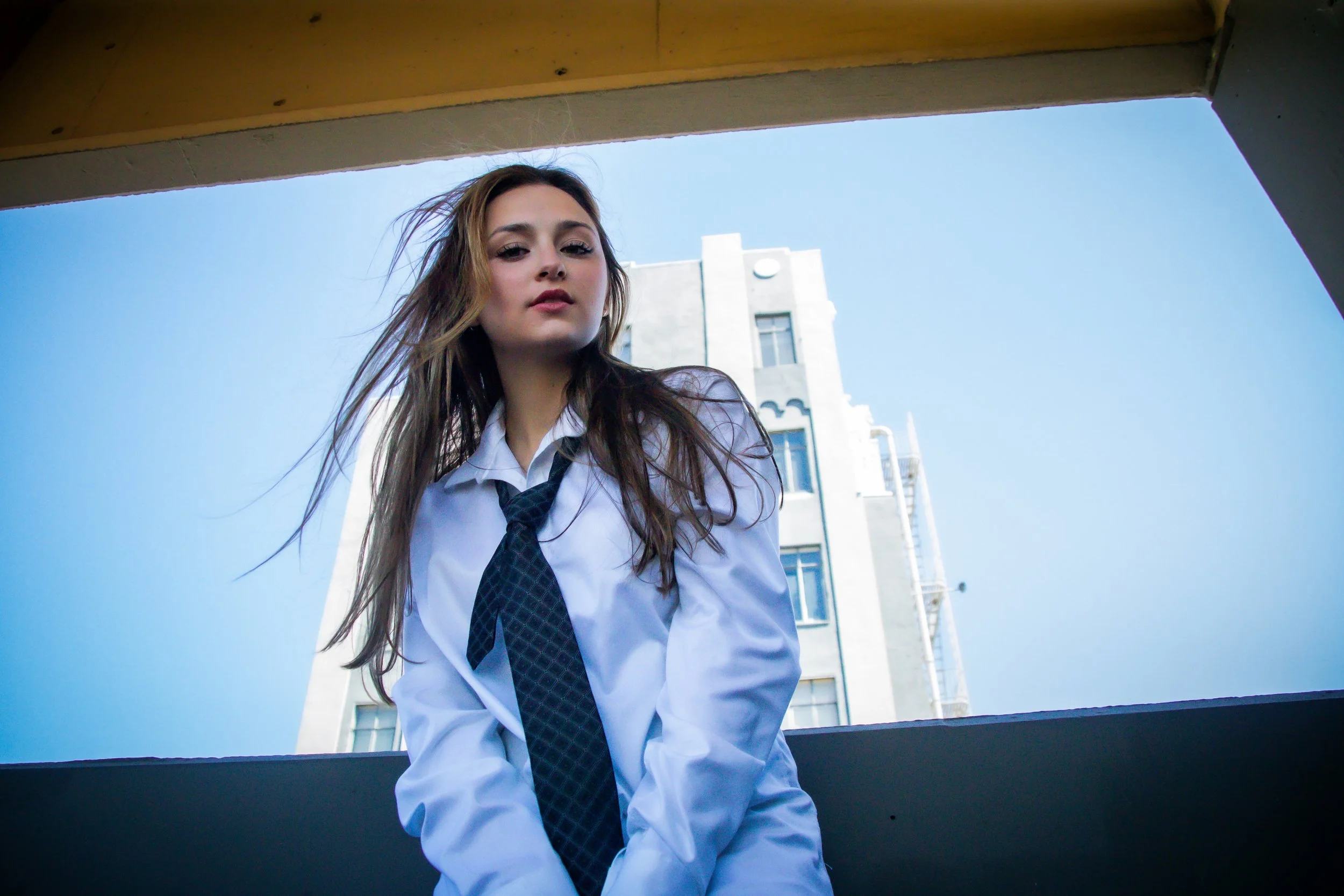 Stylish low-angle photo of a person in a white shirt and black tie standing confidently in front of a city building with a fire escape—capturing movement through wind-blown hair and a strong urban aesthetic.
