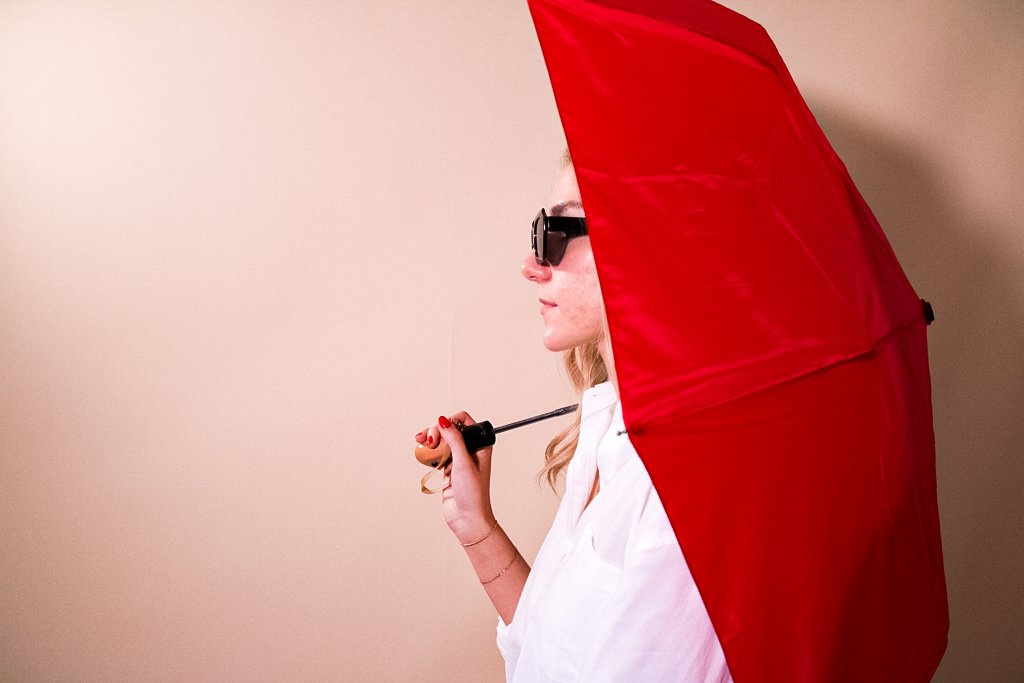 Portrait of a person in sunglasses and a white shirt holding a red umbrella against a neutral beige wall—stylized with clean lines, minimalist contrast, and modern fashion elements.