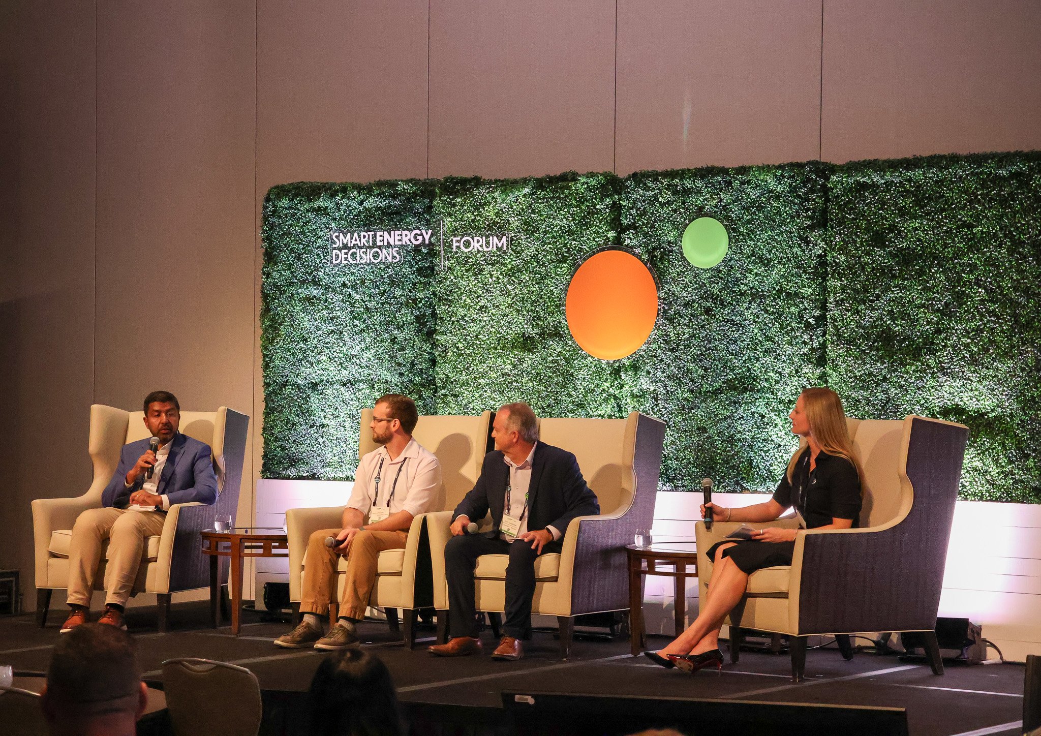 Panel discussion at the Smart Energy Decisions Forum, with four speakers on stage seated in high-backed armchairs, one person speaking into a microphone, and a green foliage backdrop with the event name.