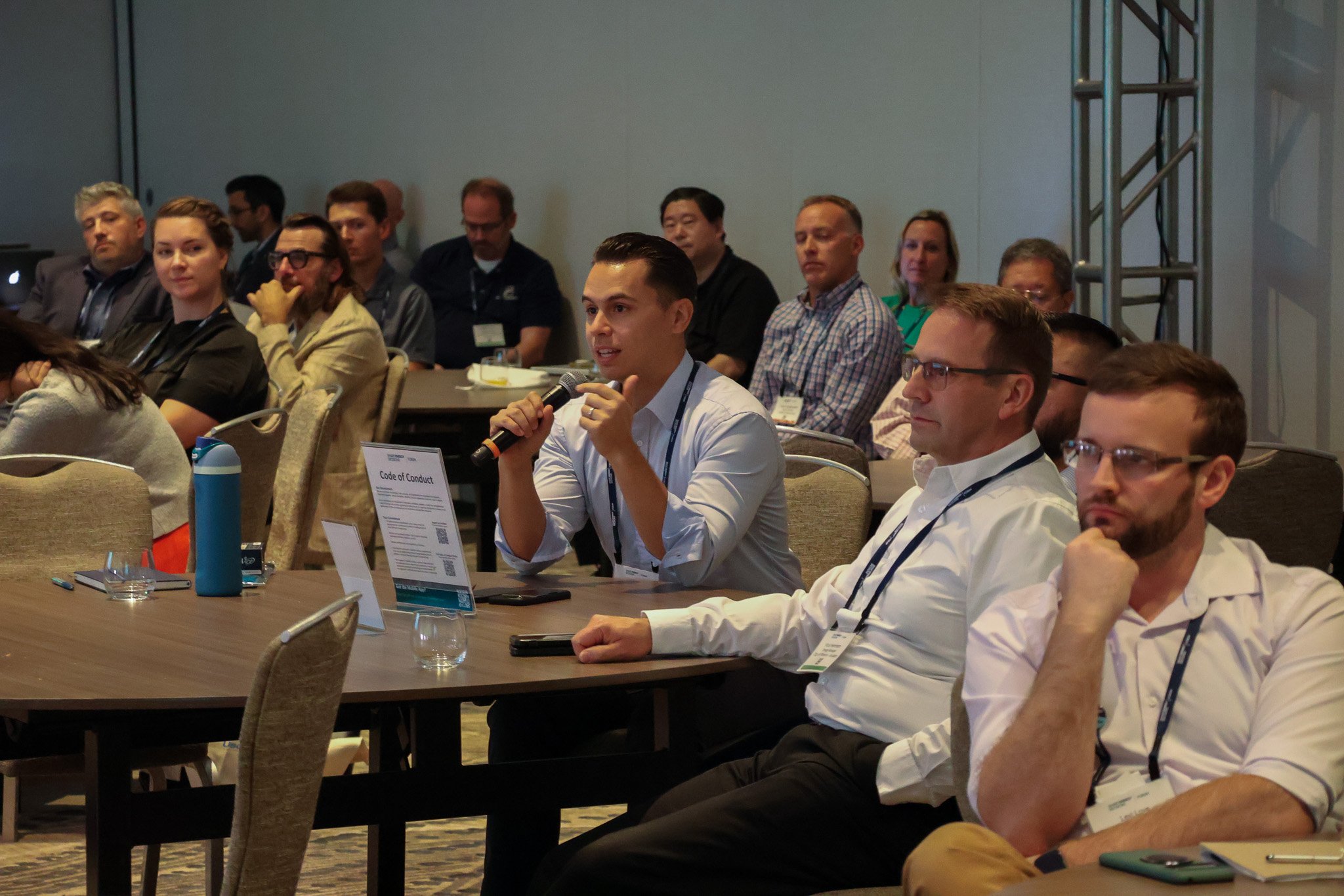 A group of people attending a conference, with one young man in a white shirt holding a microphone and speaking. Others are seated at tables with notebooks, water bottles, and conference materials.
