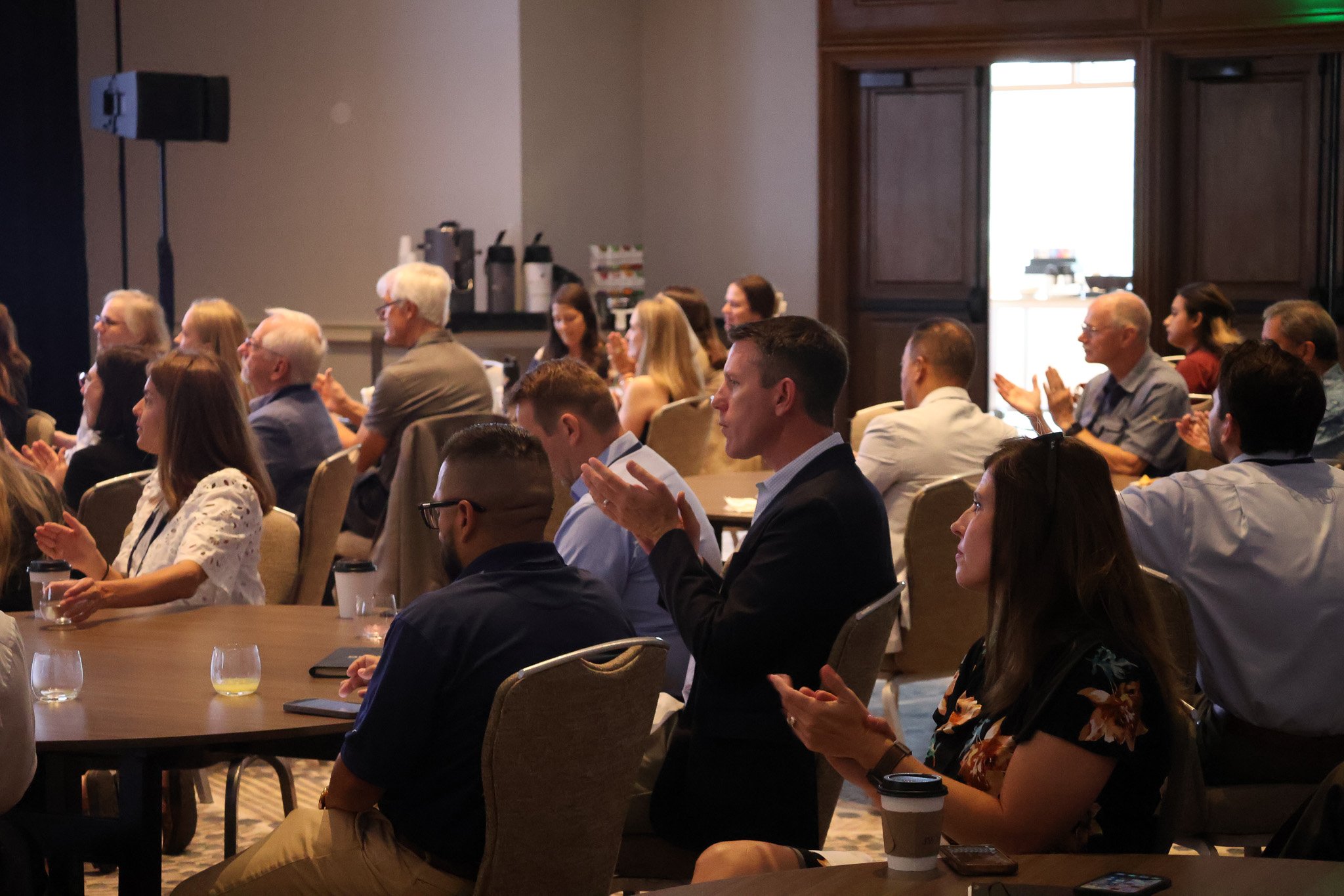 People attending a conference or seminar, seated and clapping in a dimly lit room.