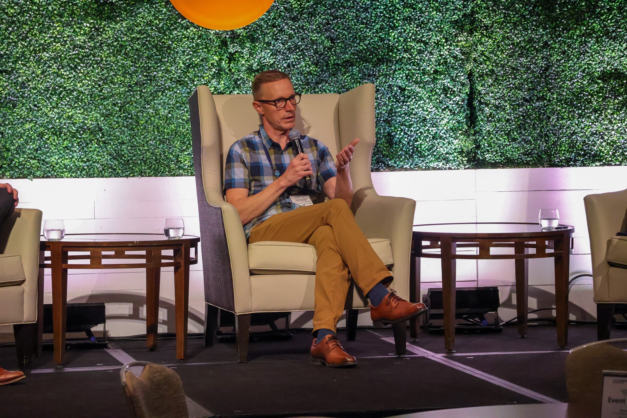 A man with glasses, wearing a plaid shirt and tan pants, sitting in a high-backed chair on a stage, speaking into a microphone, with a green leafy backdrop and side tables with water glasses.
