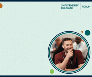 A smiling man in a maroon shirt sitting at a table during a discussion or meeting, with a graphic design background titled 'Smart Energy Decisions Forum'.