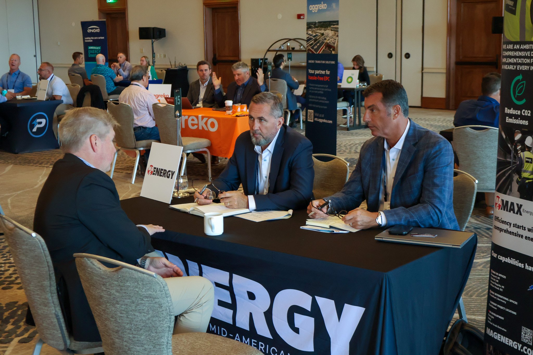 Business professionals sitting at a table during a conference or trade show, with banners and booths visible in the background.