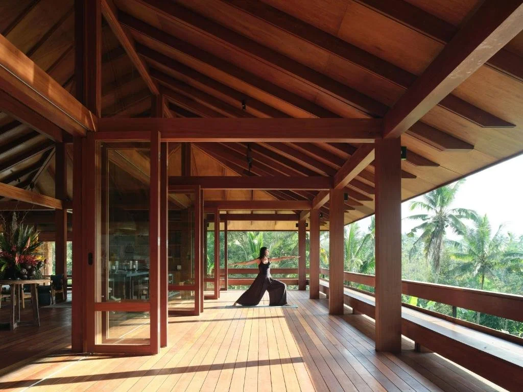 A woman practicing yoga or stretching at a luxary retreat in Bali on a wooden balcony with a natural tropical forest view, surrounded by wooden beams and open sliding glass doors.