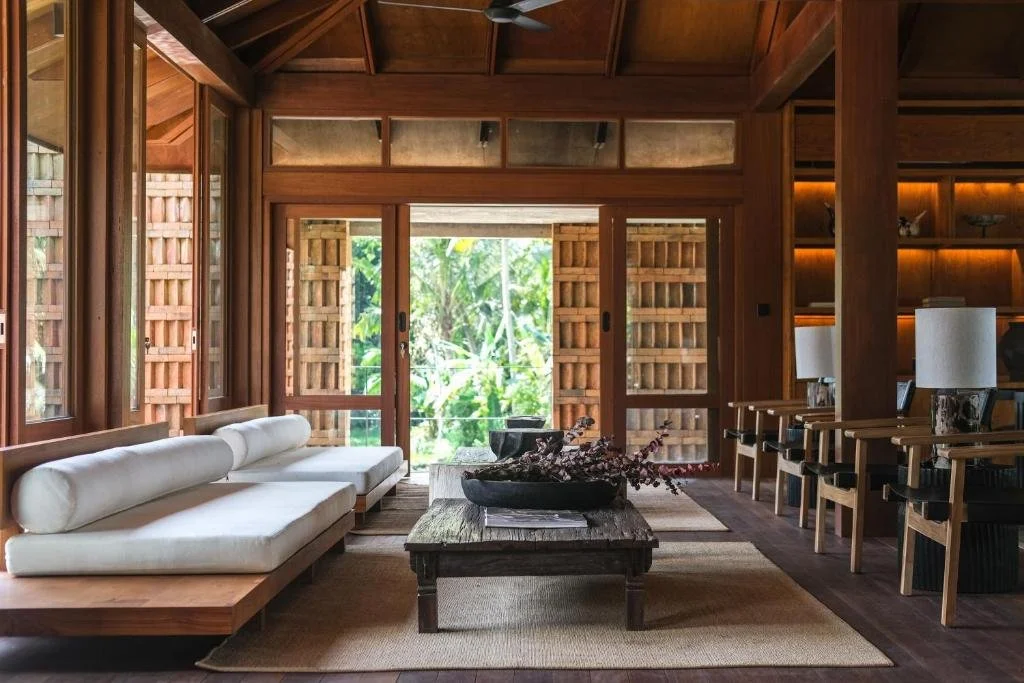 Living room with wooden walls, sliding glass doors, a white sofa, a wooden coffee table with a floral arrangement, and chairs with black cushions  at a luxary retreat in Bali.